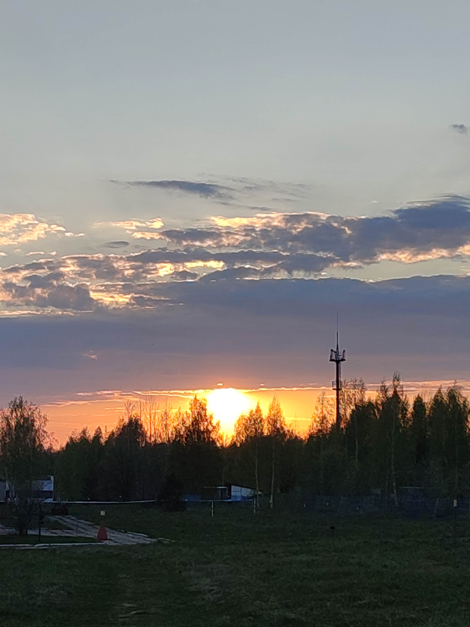 The image depicts a sunset over a landscape of trees and a grassy field. The sun is low on the horizon, partially obscured by the trees, creating a bright orange glow. A tower with antennas is visible to the right of the sun. Clouds are present in the sky, with varying shades of gray, pink, and light blue. In the foreground, there is a grassy field, and some buildings are visible in the mid-ground to the left.