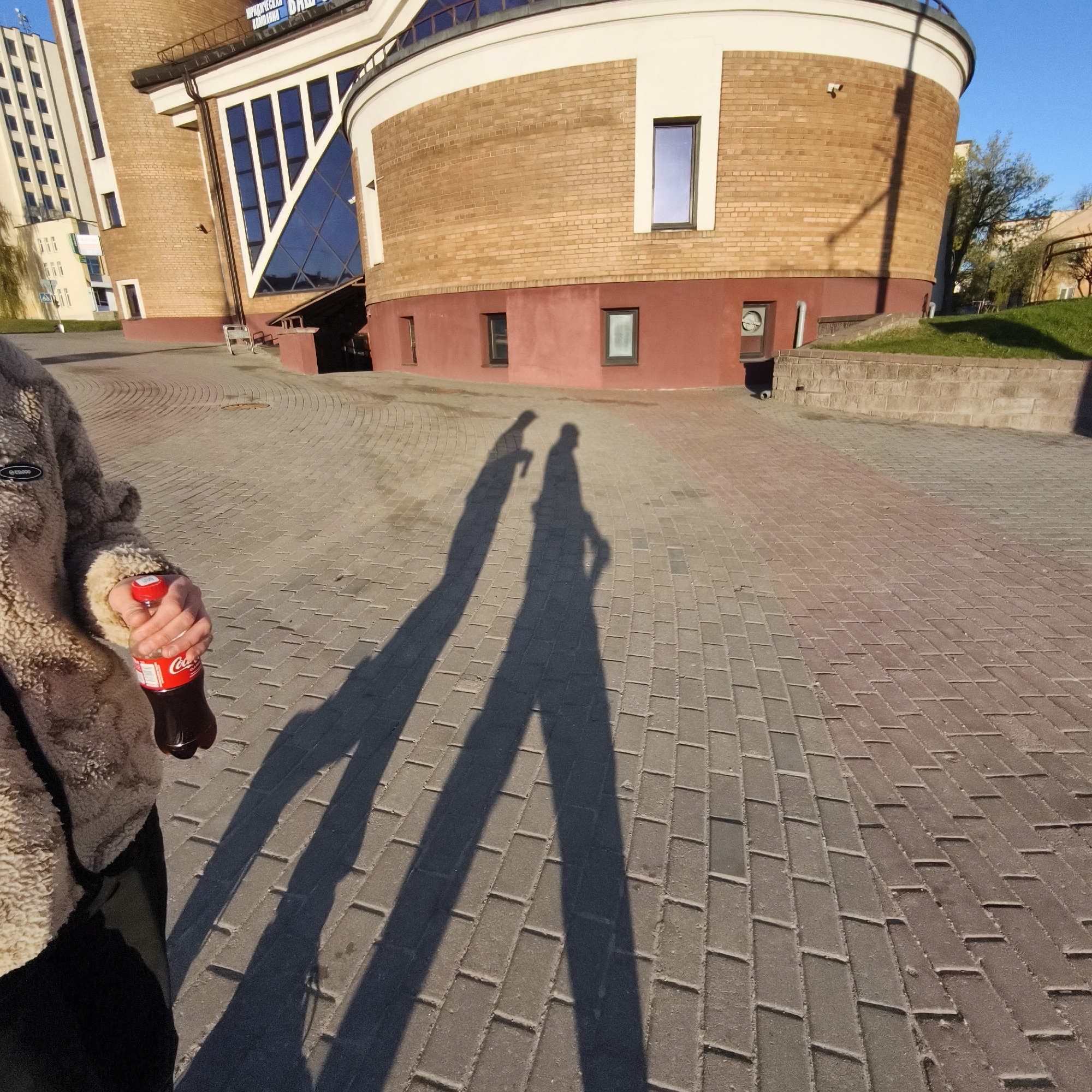The image shows a street view in Hrodna, Belarus. The shadows of two people are cast on a brick-paved walkway. The person on the left is holding a bottle of Coca-Cola. The background includes part of a brick building with a cylindrical section and a triangular window feature. Another building is visible in the distance. There is also a small patch of green grass.