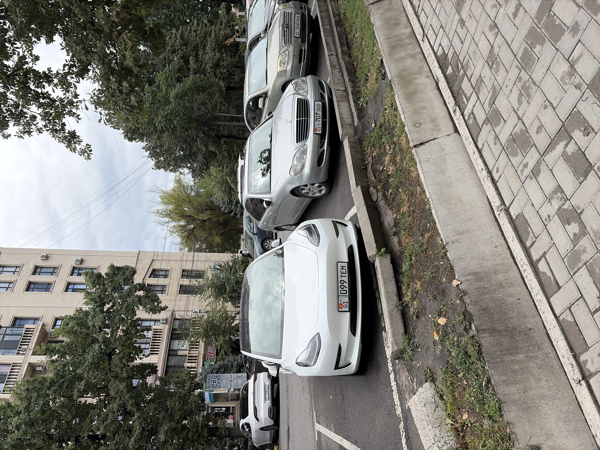 The image depicts a street scene in Bishkek, Kyrgyzstan. In the foreground, a white Tesla Model 3 is parked, facing the viewer. The car has a license plate reading "01 G99 TEM". To its right, a silver Mercedes-Benz S-Class is parked, with license plate "01 707 SUS". Further to the right, a silver Toyota Avensis is visible with a license plate "04 540 AHS". In the background, a modern apartment building stands tall, with trees and power lines in front of it. The street is paved with cobblestones, and a concrete curb runs along the side. The sky is overcast, suggesting a cloudy day. No people are visible in the image. The overall scene is one of urban street parking.