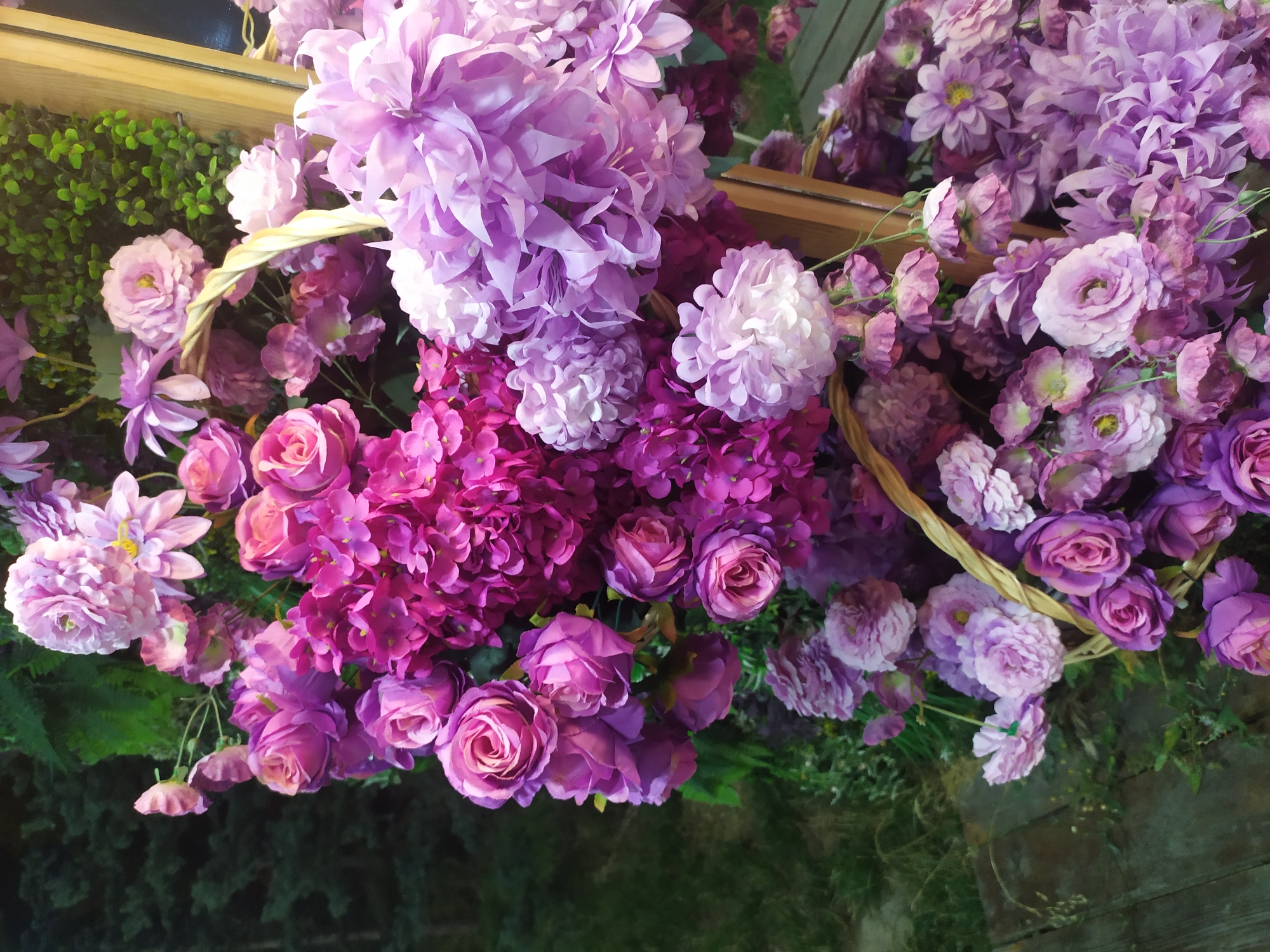 A close-up, high-angle shot features a lush display of artificial flowers in various shades of purple and pink. The flowers are arranged in a cascading fashion, spilling out of woven baskets and against a backdrop of dark green foliage. The textures vary from the tightly packed petals of hydrangeas and roses to the more open, layered petals of chrysanthemums and dahlias. The lighting appears to be ambient, creating soft shadows and highlighting the different colors and forms of the blooms. The overall impression is one of abundance and decorative artistry. The image does not provide any specific identifiable landmarks or features that would definitively place it within the city of Sosnovka, Kyrgyzstan.