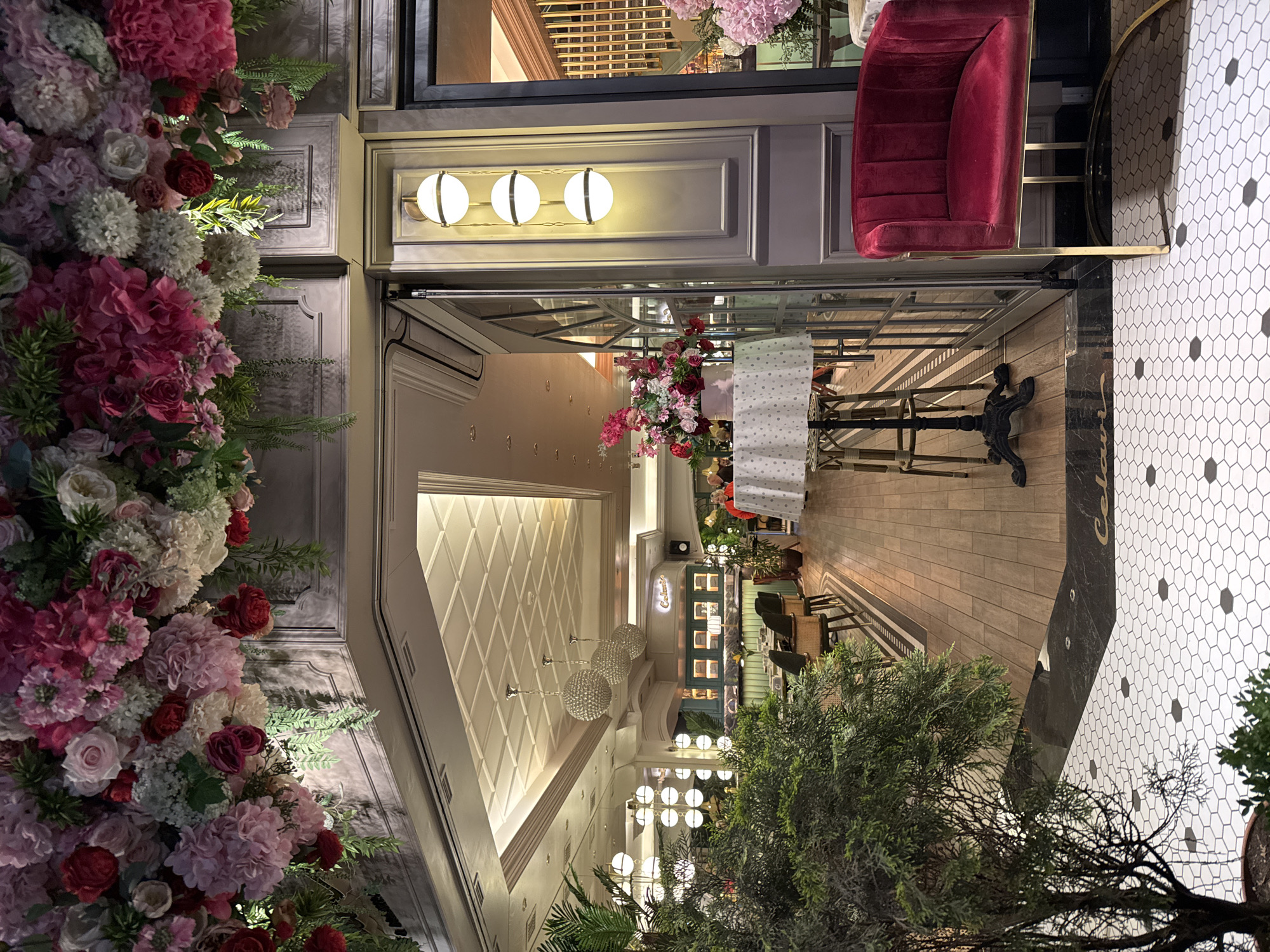 The image shows the interior of a restaurant in Yerevan, Armenia. The entryway is adorned with a profusion of pink and white flowers and lush greenery. Three globe-shaped lights illuminate the entrance area, above a door leading into a dining space. The dining area features wooden floors, tables set for service, and chandeliers hanging from a decorative, geometrically patterned ceiling. A red velvet chair is positioned near the entrance, beside a floor covered with white hexagonal tiles with black accents. The overall ambiance appears elegant and refined. Outside, more greenery frames the view.