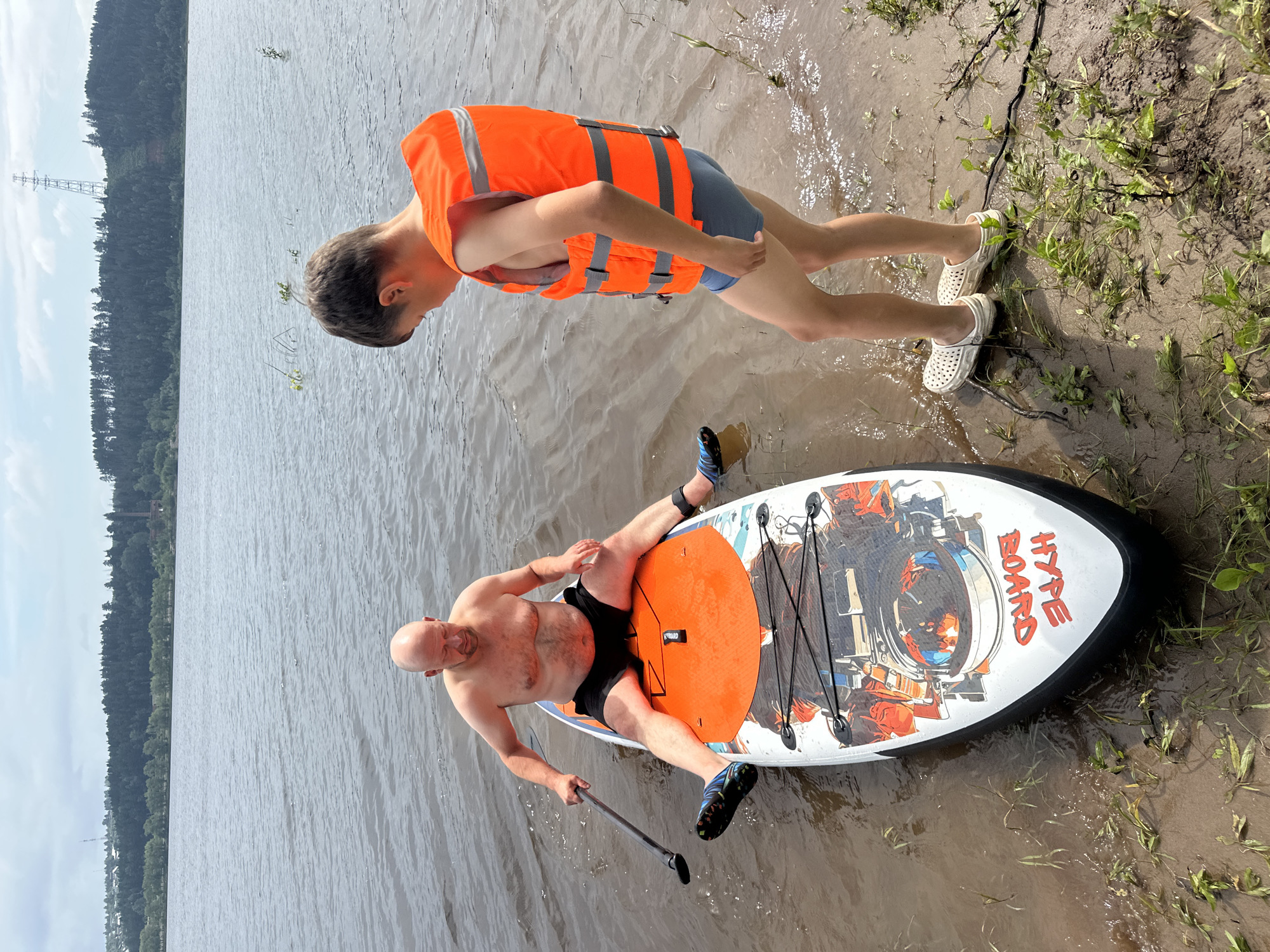 A bald, shirtless man wearing black shorts sits on an orange and white paddleboard in shallow, muddy water near the shore of the Kama River in Chaikovsky, Russia. He holds a paddle in his right hand. A young boy wearing an orange life vest and blue swim trunks stands on the muddy shore to the right of the paddleboard, looking down. The background shows a dense forest on a hillside and a distant power line tower. The sky is overcast.