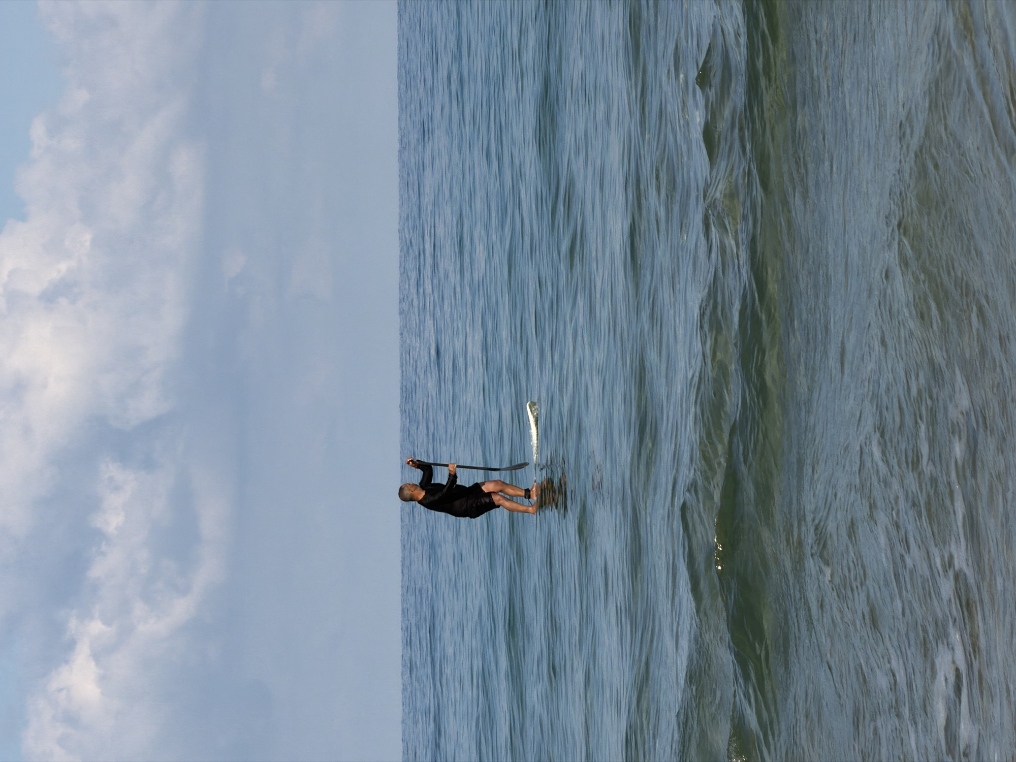 The image depicts a man stand-up paddleboarding on the sea, likely in Bat Yam, Israel, based on the environment. The man is wearing a black wetsuit and is holding a paddle, standing on a board that is moving through the water. The right side of the image shows choppy waves, while the left side depicts a clear sky with white clouds.