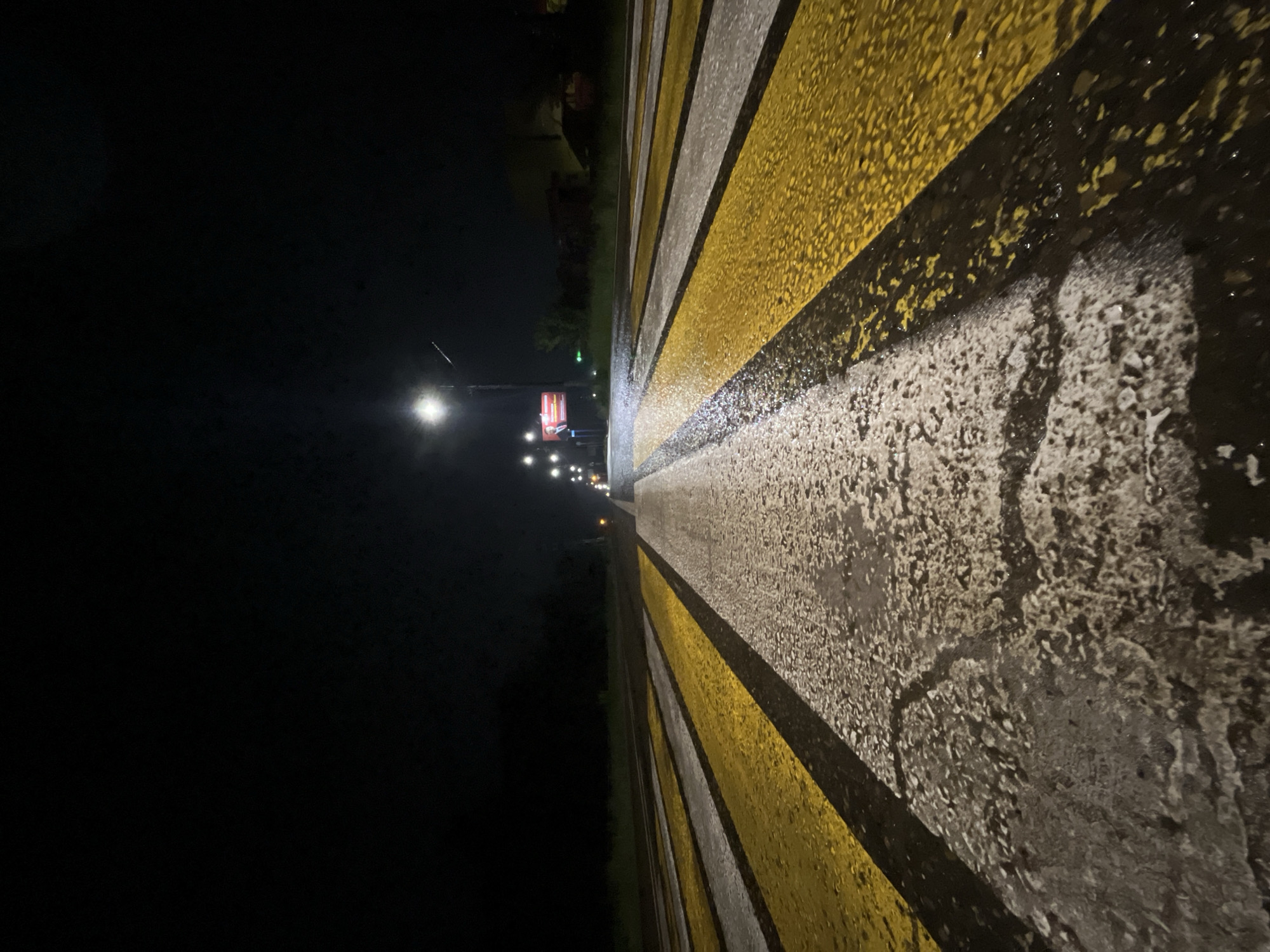 A low-angle forensic photograph depicts a wet pedestrian crossing on an asphalt road at night in Chaikovsky, Russia. The foreground shows a detailed view of the road surface, which is covered in a film of water, causing the white and bright yellow striped markings of the crosswalk to appear highly reflective and textured. These stripes extend from the bottom of the frame, converging towards the distant horizon.
The environment is dimly lit by artificial sources. A prominent streetlight on the right side of the road casts a bright, localized glow. Further along the right side, a series of additional streetlights illuminate the roadway and a narrow strip of vegetation or faint structures. In the mid-distance on the right, a rectangular billboard, brightly illuminated, displays a graphic with an image of a man in a suit and visible text. The left side of the road recedes into deep darkness, with only vague, dark silhouettes suggesting trees or undeveloped land. No individuals, vehicles, or explicit actions are present in the scene, which conveys a sense of stillness on a wet, artificially lit urban road.