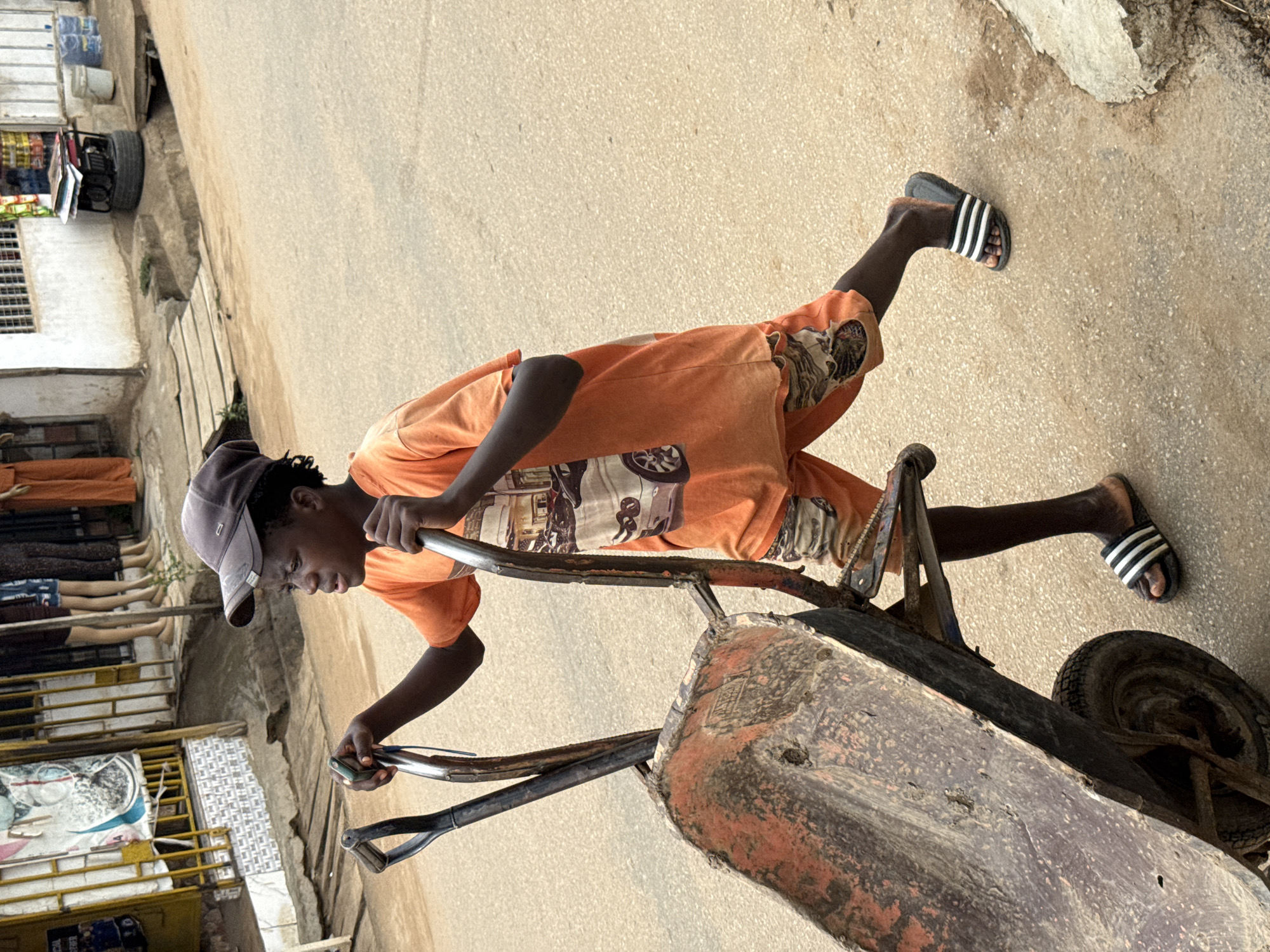 The image captures a young person, likely male, pushing an old, rusty wheelbarrow down a paved street in what appears to be an urban or semi-urban setting. He is dressed in an orange t-shirt, orange patterned shorts, a dark baseball cap, and black-and-white striped slides. His expression is focused, suggesting engagement in a task or labor. The wheelbarrow itself shows significant wear and tear, with rust and dirt evident on its metal frame and bucket.
In the background, on the left side of the frame, there are several structures that resemble shops or stalls. One stall displays what appear to be bottled drinks, and below it, mannequin legs are visible, suggesting a clothing display. Further back and higher up, a generator or similar piece of machinery is visible. The buildings have concrete walls, some with barred windows. The ground is a light-colored, granular surface, likely asphalt or concrete.
The lighting indicates daytime, possibly mid-morning or afternoon, with soft shadows suggesting clear weather. The overall scene depicts a typical street view, likely in a bustling area. No discernible text is clearly visible on any signs, clothing, or objects within the image. The visual cues, such as the architecture, street vendors, and general atmosphere, are consistent with the provided location context of Abuja, Nigeria.