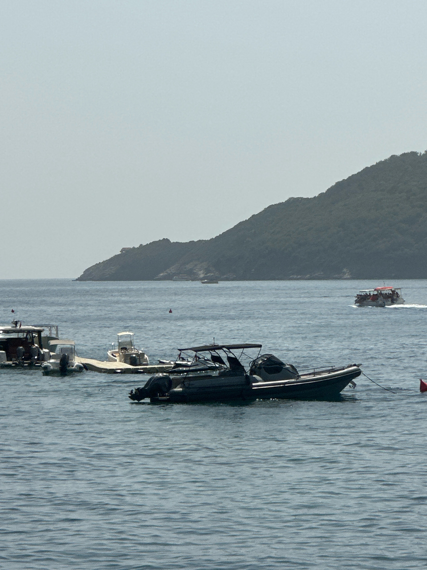 A fleet of boats is moored in the calm waters of Budva, Montenegro, with a verdant, tree-covered hill rising in the background. In the foreground, several smaller boats are docked, including a dark-colored inflatable boat with a black canopy and an outboard motor. To the left, a larger white boat with a cabin is partially visible, with people standing on its deck. Further in the distance, a tour boat filled with passengers is cruising on the water, leaving a wake. The sky is a hazy light blue, and the overall scene suggests a peaceful day in a coastal town.