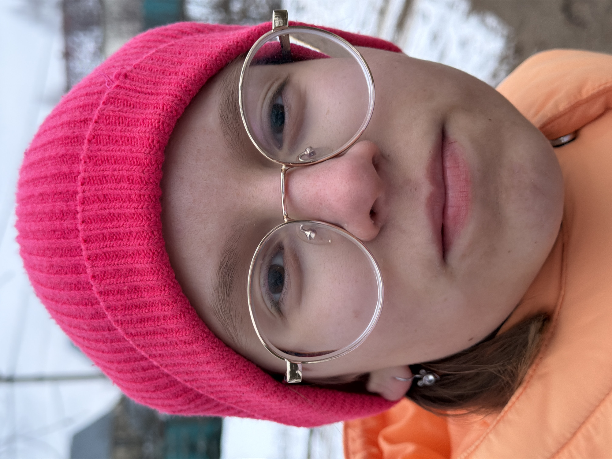 A close-up portrait shows a person wearing a bright pink knitted beanie and round, gold-framed glasses. The person is also wearing an orange jacket. The background is slightly blurred, but suggests an outdoor setting with hints of snow or a light-colored building. There is no specific information to confirm the location is in Chaikovsky, Russia.