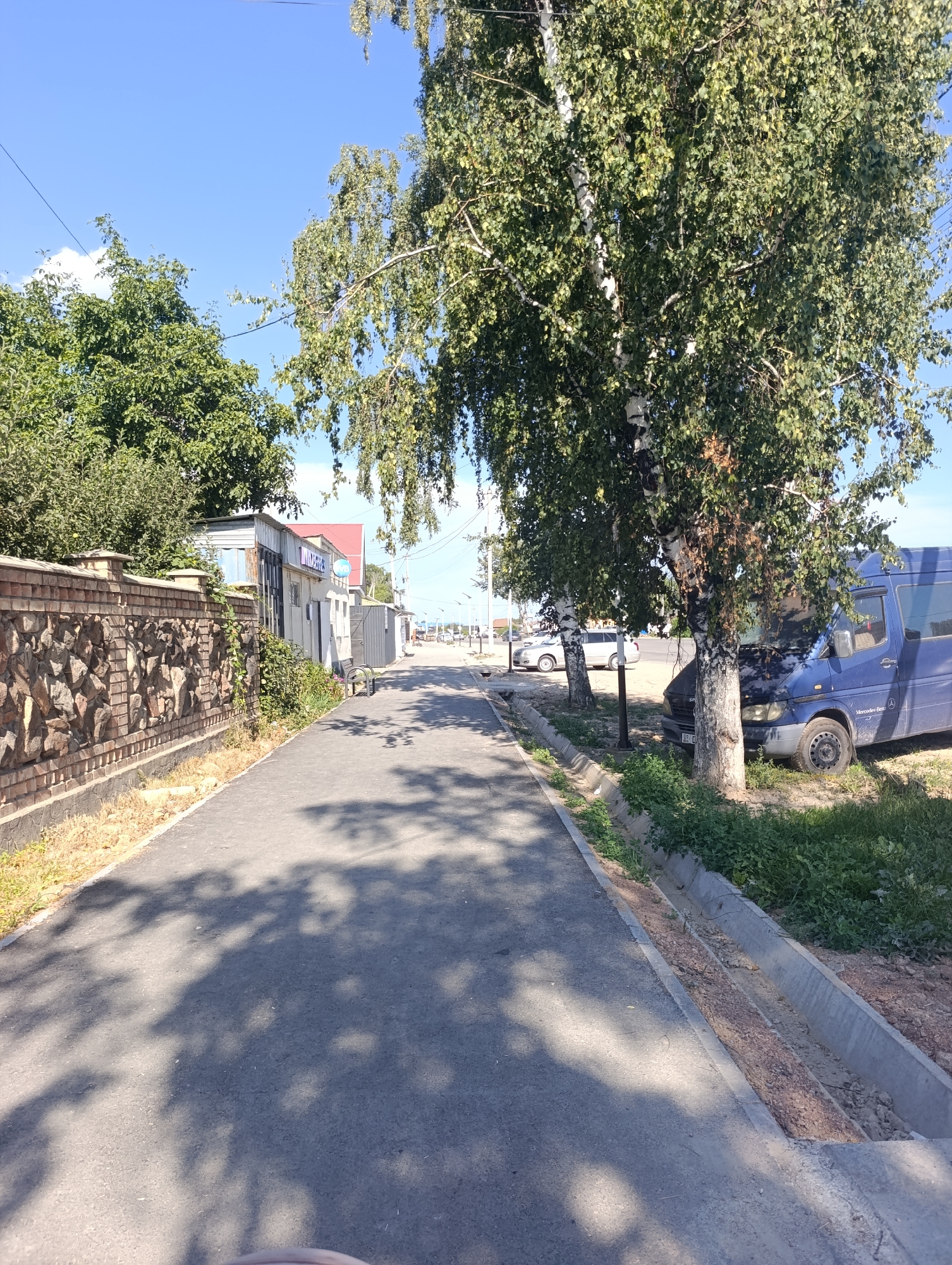 The image depicts a sunny day in Bosteri, Kyrgyzstan, showing a paved walkway next to a low stone wall. Beyond the wall are buildings, one of which has signage that appears to be in Cyrillic. Several trees line the walkway, casting shadows on the ground. A blue van is parked to the right of the walkway, partially obscured by trees. Further down the walkway, other vehicles and buildings are visible, suggesting a commercial or residential area. The sky is clear and blue.