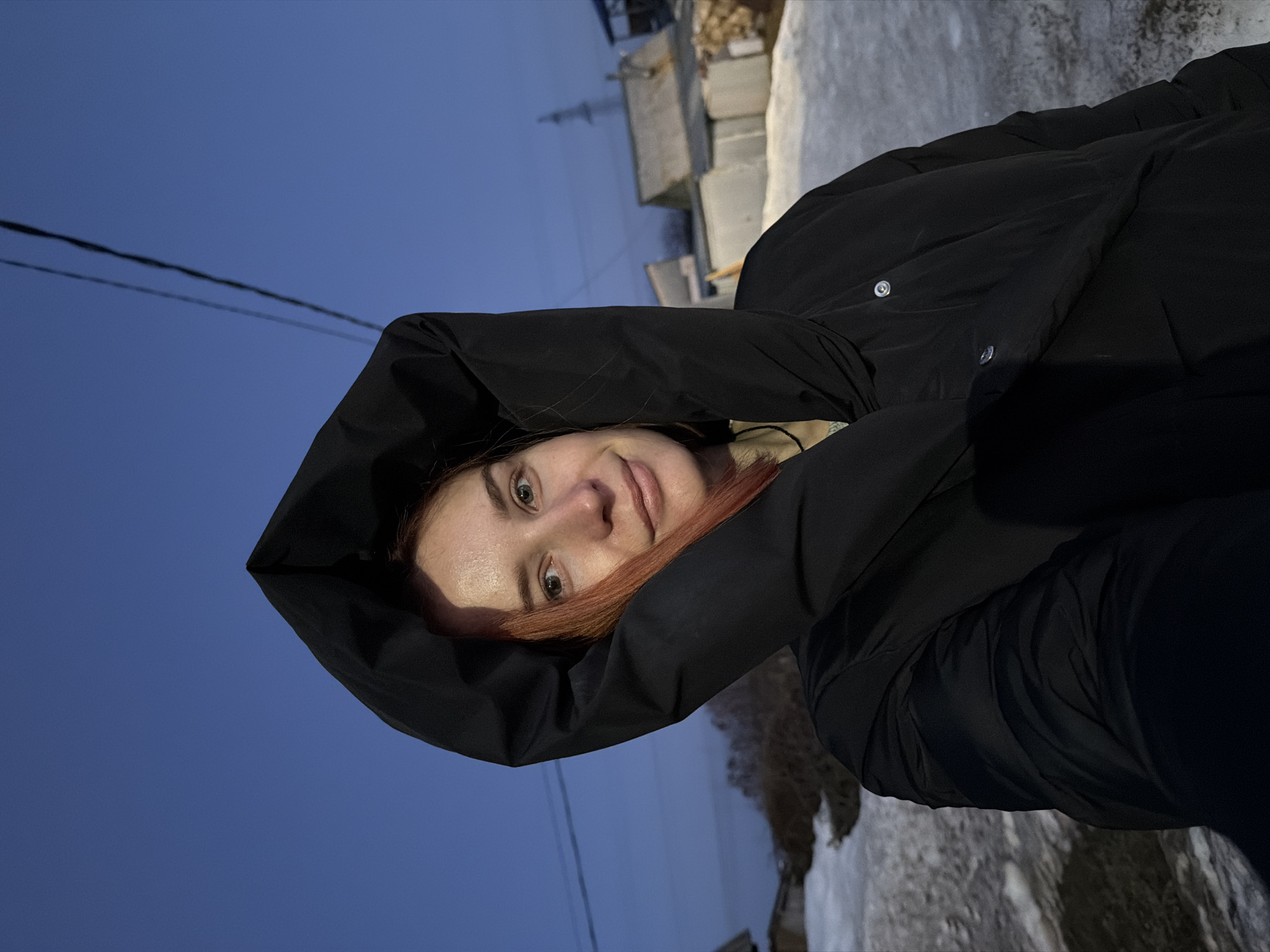 A woman with fair skin and reddish-orange hair is wearing a black, puffy jacket with the hood up. She is facing the camera with a slight smile. The background shows a blue sky transitioning to dusk or night. There are utility wires visible in the sky. Behind her, there appear to be buildings or structures constructed from cinder blocks and patches of snow on the ground. The image is rotated counterclockwise, and based on the background details, the location appears to be Chaikovsky, Russia.