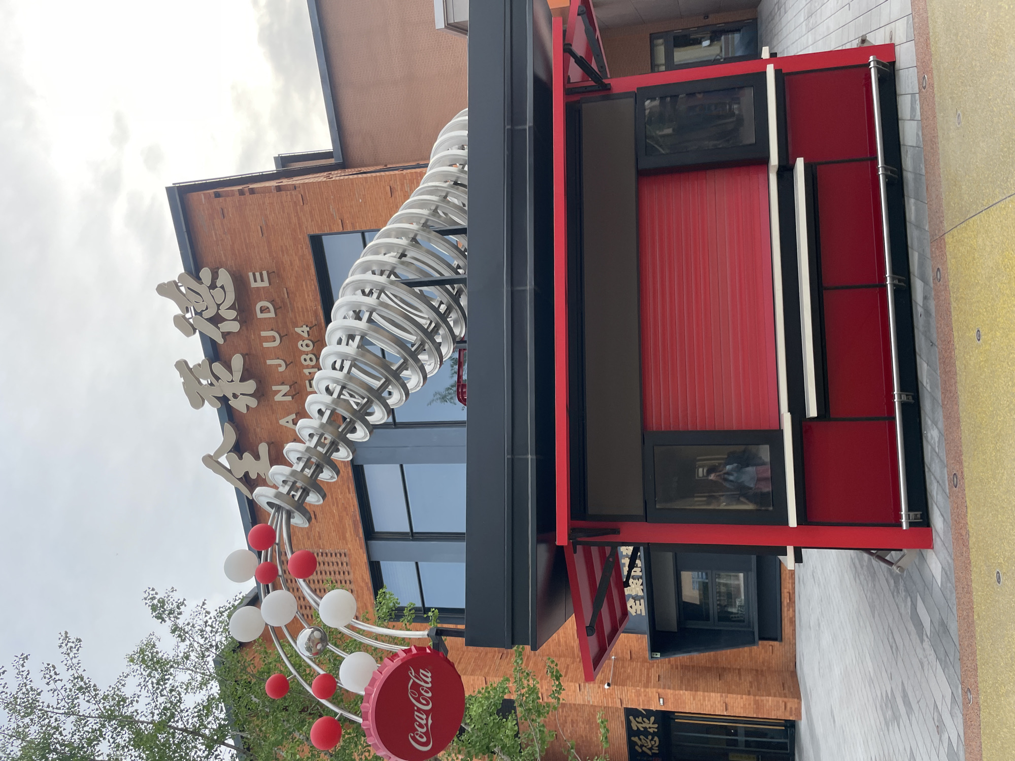 The image captures a street scene in Tongzhou District, China, showcasing a vibrant blend of modern commercial elements and architectural design. A red brick building serves as the backdrop for a Coca-Cola-themed stall or small store, distinguished by a large Coca-Cola bottle cap logo and red and white decorative balloons. A metallic, coiled structure adds a sculptural touch to the scene, positioned adjacent to Chinese characters and the inscription "NJUDE 1864," possibly indicating a historic connection or branding element. The store's façade features a combination of black and red panels, with a red shuttered section and open windows, and some reflective windows with people visible in the reflection. The street is paved and there is some greenery.