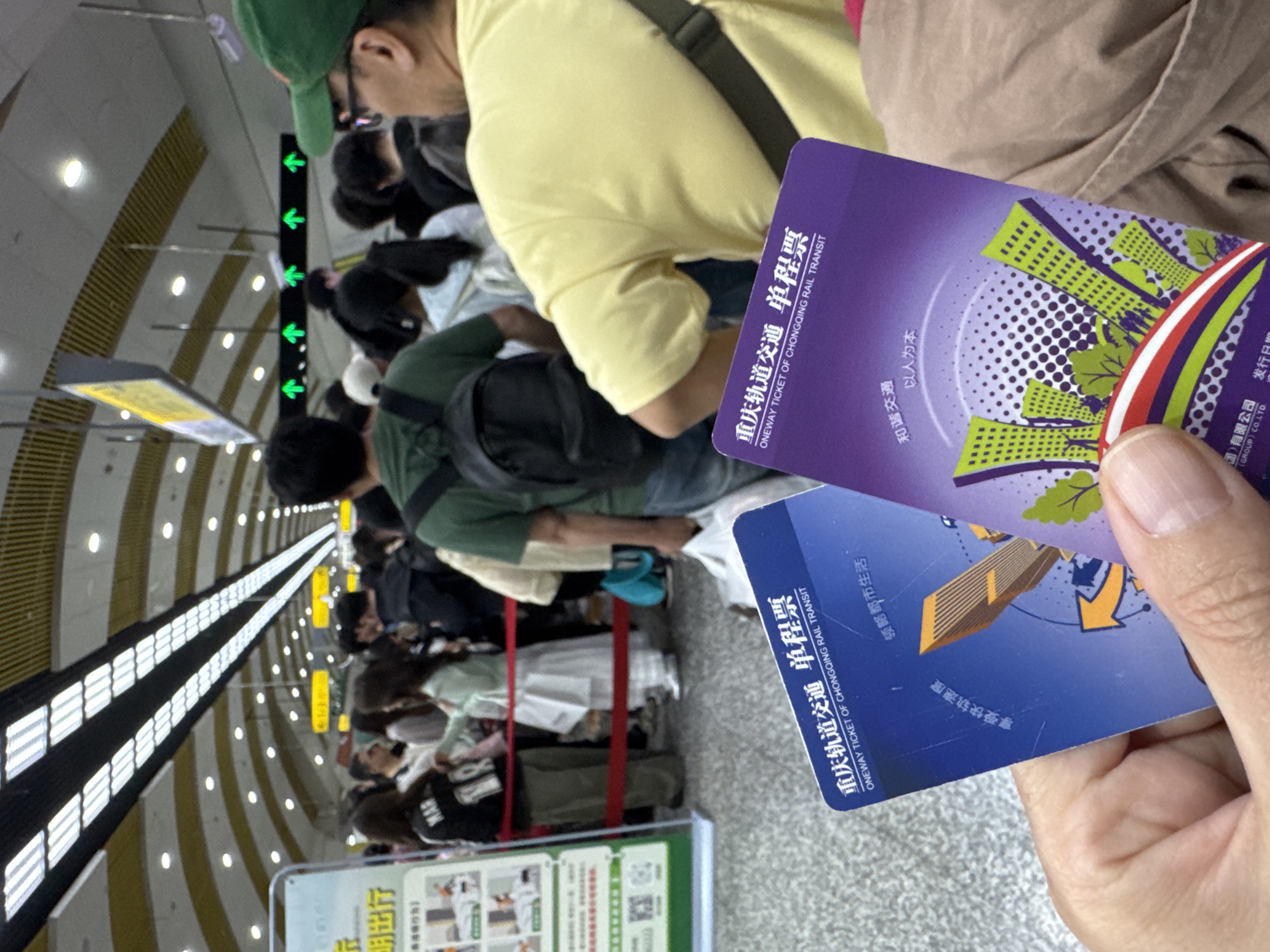 The image shows a first-person view of two transit tickets for the Chongqing Rail Transit system held in a hand. The tickets are for a one-way fare. The purple ticket on top features a cityscape graphic with green and yellow elements. The blue ticket has a similar design, depicting a tall building with orange arrows. The English text on the tickets reads "ONEWAY TICKET OF CHONGQING RAIL TRANSIT".
In the blurred background, there is a line of people, possibly waiting in a queue or on a train platform. There are directional signs with green arrows, overhead lighting, and a display with text in Chinese. The scene appears to be inside a station, possibly in the Yuzhong District of Chongqing, China, given the information on the tickets.