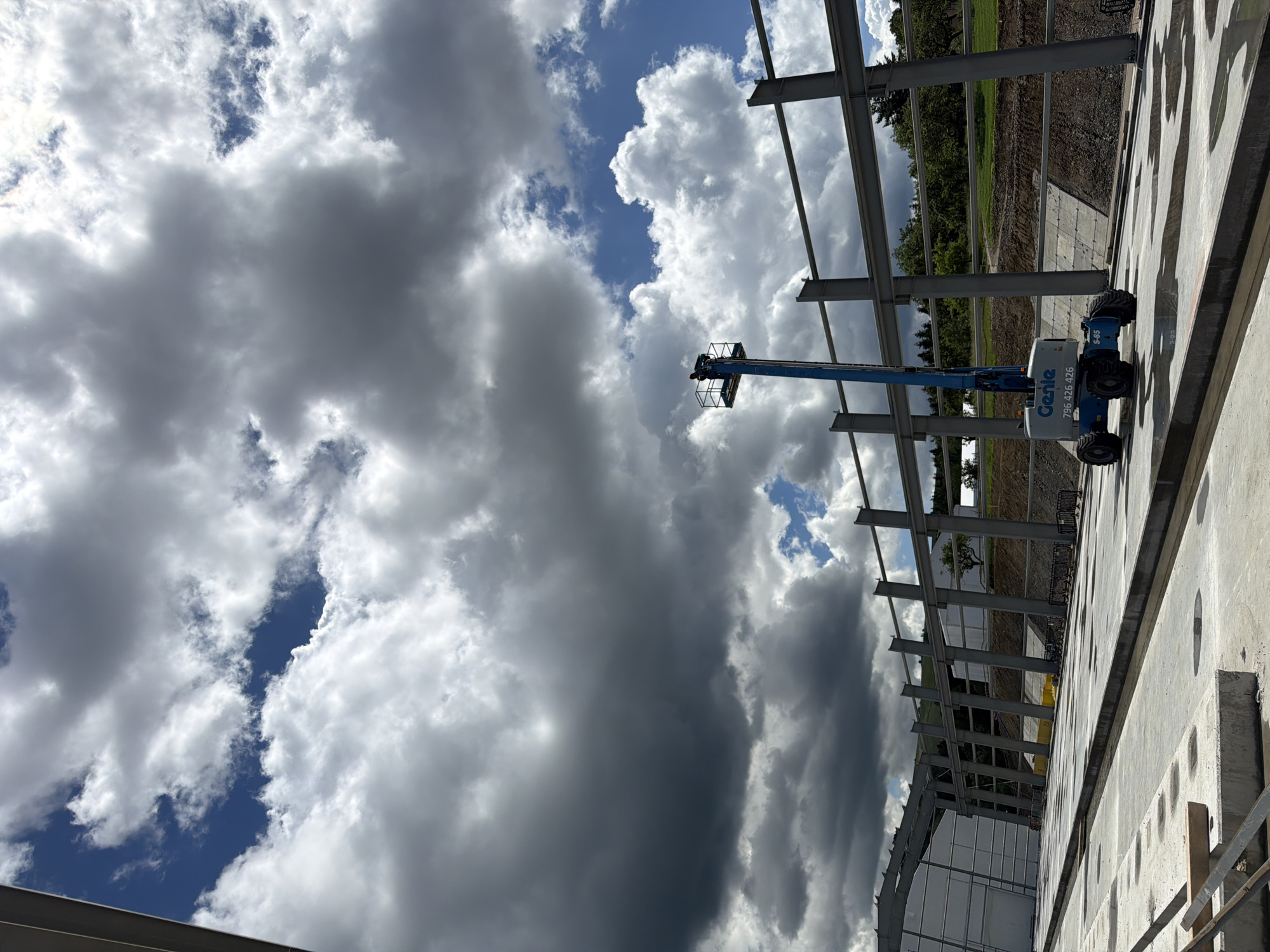 In Janowice, Poland, a construction site is captured under a dramatic sky filled with dark, heavy clouds interspersed with patches of blue. A blue Genie aerial work platform, with the model number S-85 visible, is positioned on a wet concrete surface. The platform is extended, and a person can be seen in the basket at the top. In the background, a large steel-framed structure is under construction, with rows of structural beams and columns visible. The ground is a mix of concrete, exposed soil, and gravel. Green trees and rolling hills are visible beyond the construction area.