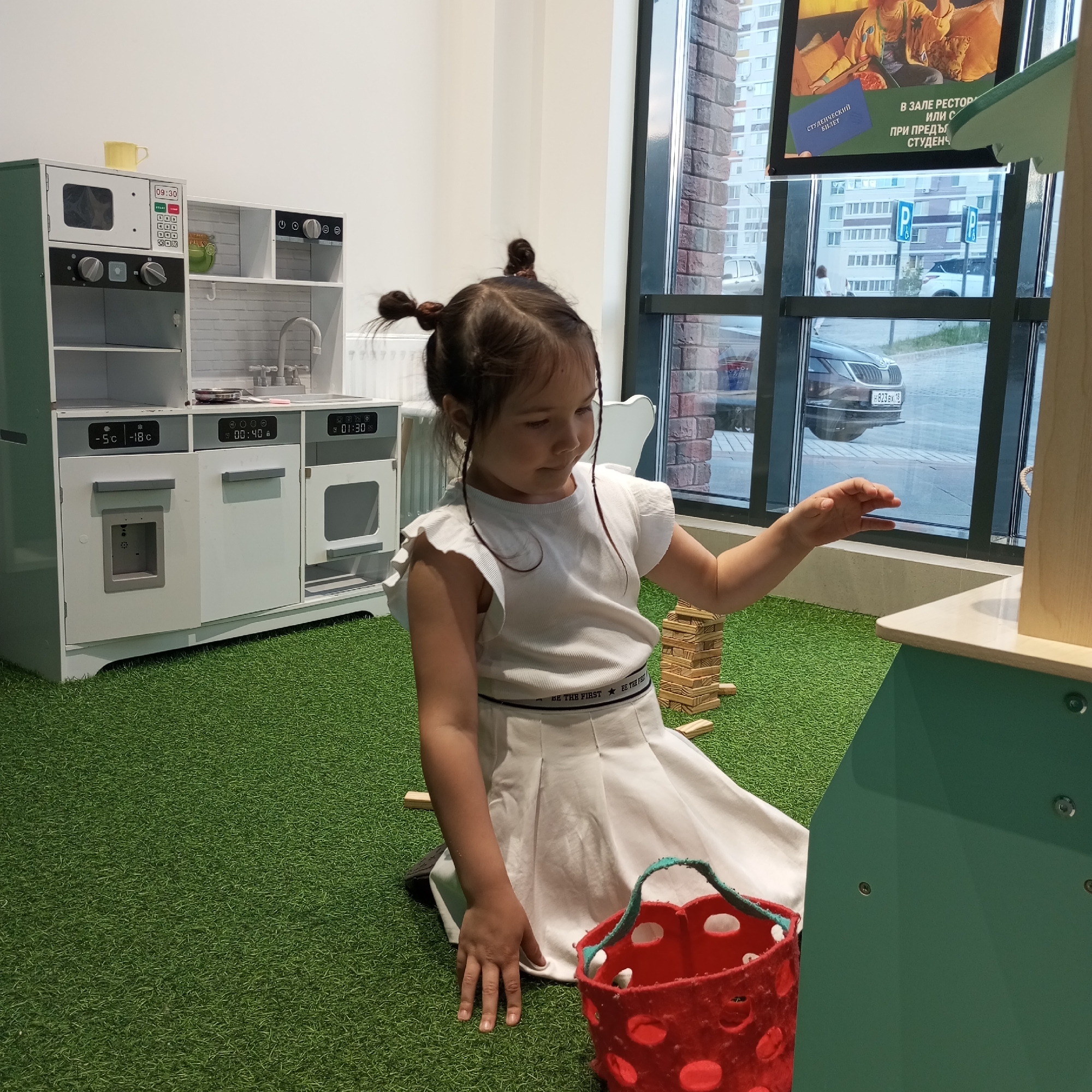 A young girl with dark hair in two pigtails is playing on a bright green artificial turf floor, indoors. She is wearing a white sleeveless top with ruffled sleeves and a white pleated skirt. She is sitting on the floor and reaching out with her right hand towards a game of Jenga blocks that is partially visible. A red basket with white polka dots is in front of her on the grass. Behind her is a white and green toy kitchen set with appliances like a microwave, oven, and sink. The kitchen set has digital displays showing "09:30", "-5C -18C", "00:40", and "01:30". Through a large window behind the kitchen set, a street scene is visible, including buildings, parked cars, and a sign for parking. The lighting suggests it is daytime.