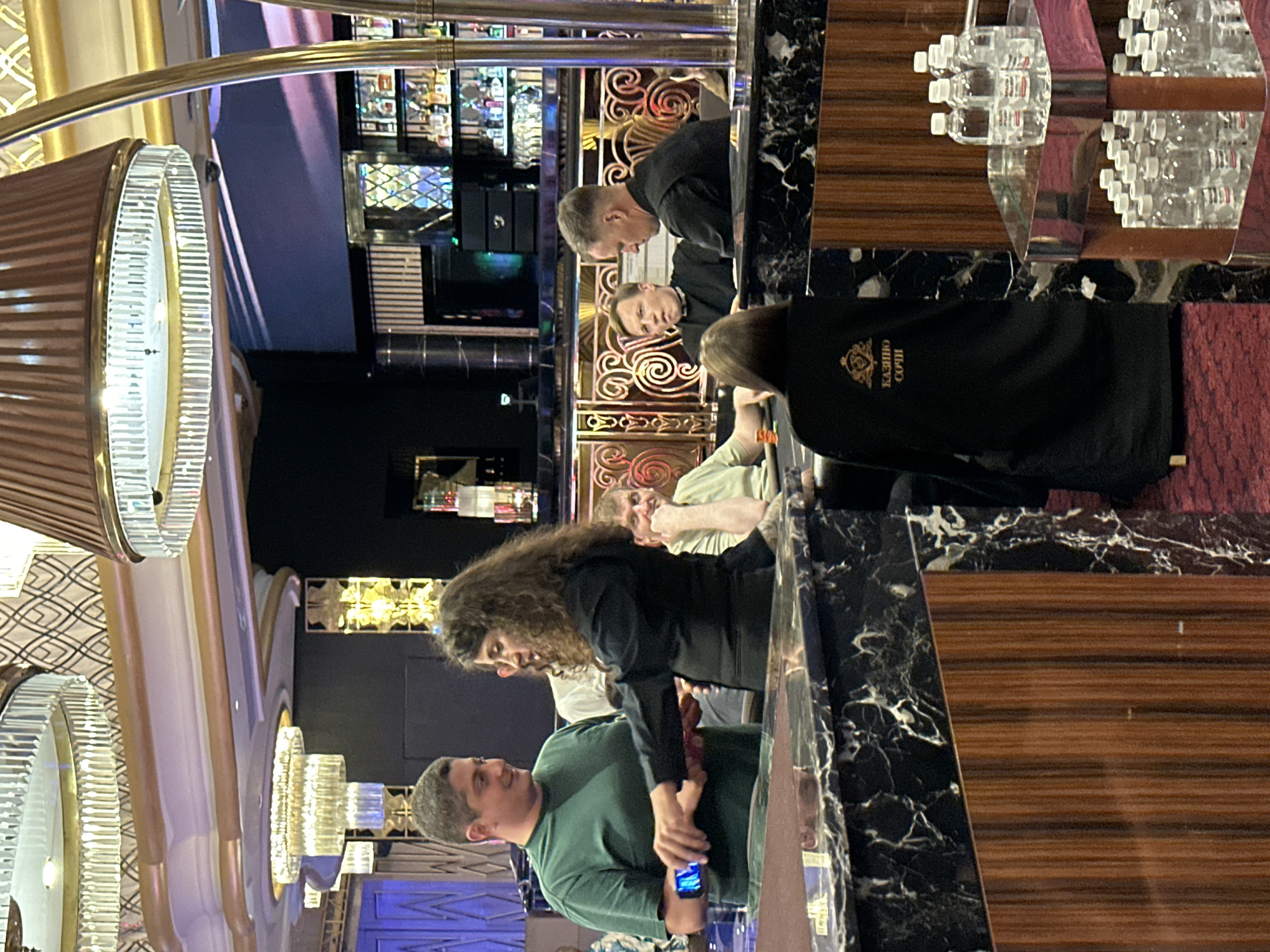 The image depicts an indoor scene at a casino, likely in Estosadok, Russia. The foreground is dominated by a dark, marbled table and a wooden paneled structure. In the middle ground, several individuals are gathered around a gaming table. On the left, a man in a green t-shirt is holding a smartphone. To his right, a woman with curly dark hair is interacting with him. Further back, other patrons are seated and appear engaged in a game. The background reveals a well-lit bar area with shelves stocked with bottles, and ornate ceiling fixtures. The overall atmosphere suggests a lively casino environment. The presence of the text "КАЗИНО СОЧИ" on the black garment worn by one of the individuals confirms the location as Sochi Casino, which is situated in Estosadok, Russia.