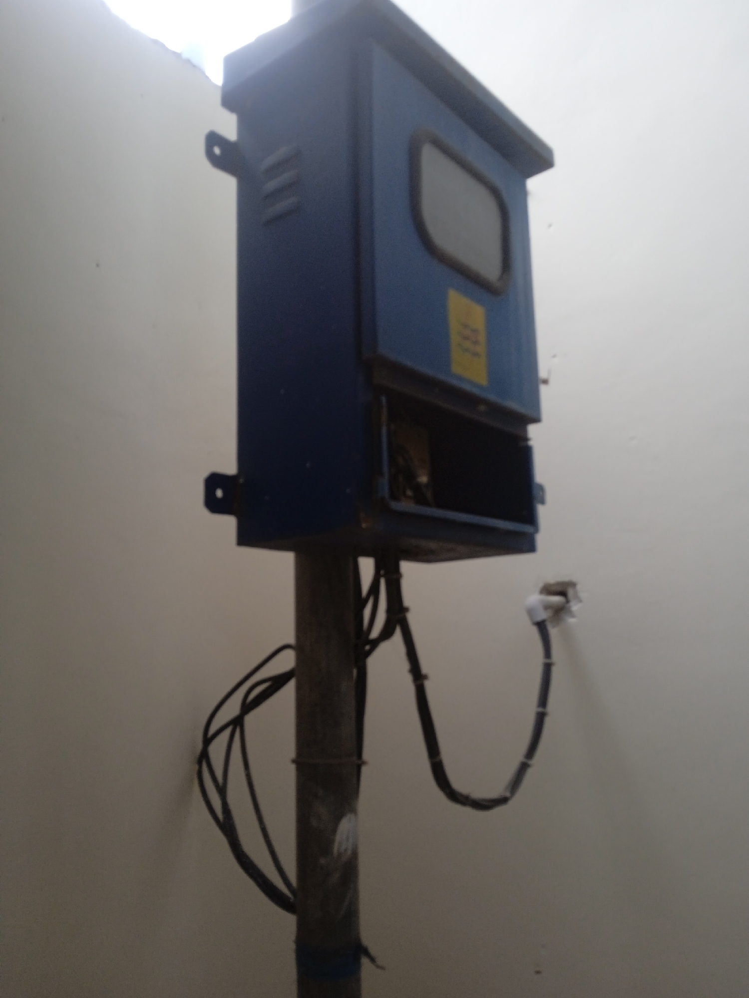 In the city of Slorok, Indonesia, a dark blue electrical cabinet is mounted on a wooden pole, positioned against a light-colored, textured wall. The cabinet has a rectangular window with a dark frame, and a yellow label is affixed below it. The cabinet door is open, revealing the dark interior. Black electrical wires hang loosely from the cabinet and are coiled around the pole. A white conduit pipe is also attached to the wall near the cabinet. The overall scene suggests an outdoor utility or junction box.