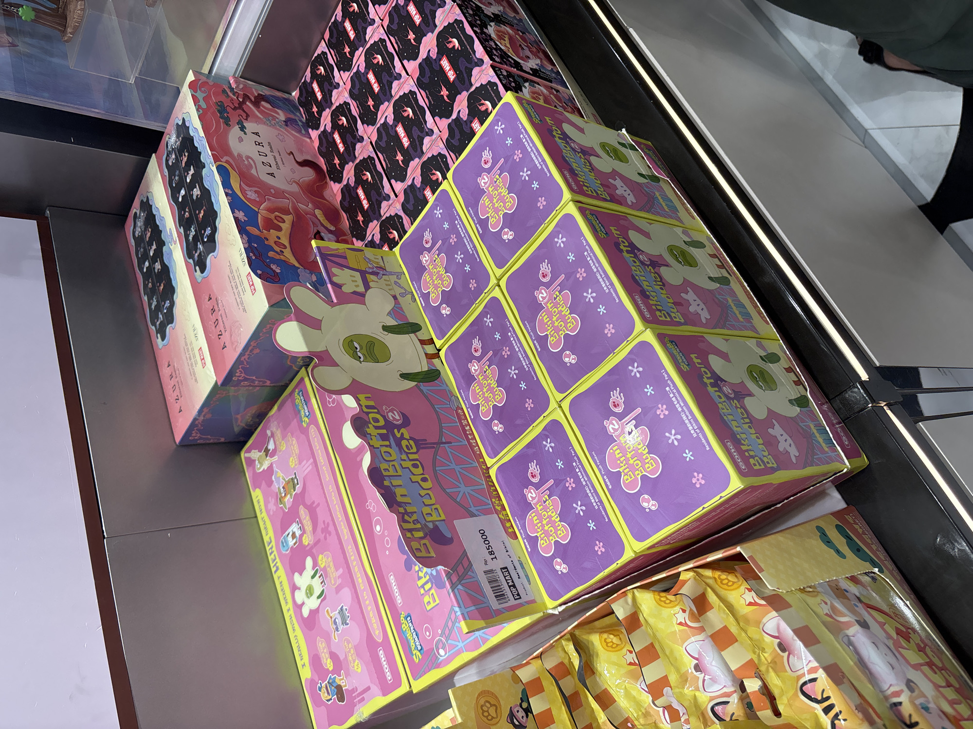 The image shows a selection of collectible toy boxes on a shelf, likely in a retail setting. The boxes are densely packed, with varying designs and color schemes. Prominently featured are boxes of "Bikini Bottom Buddies 2" in bright pink and purple packaging with cartoon characters, and a stack of "AZURA" boxes, in pink and blue. The environment appears to be a brightly lit commercial space, with a glimpse of a person's leg suggesting that the photo was taken by someone standing nearby. The barcode on one of the "Bikini Bottom Buddies 2" boxes is printed with "POP MART", which is a company with stores in malls across West Jakarta, Indonesia, but there is no certainty on whether this is the location of the photo.