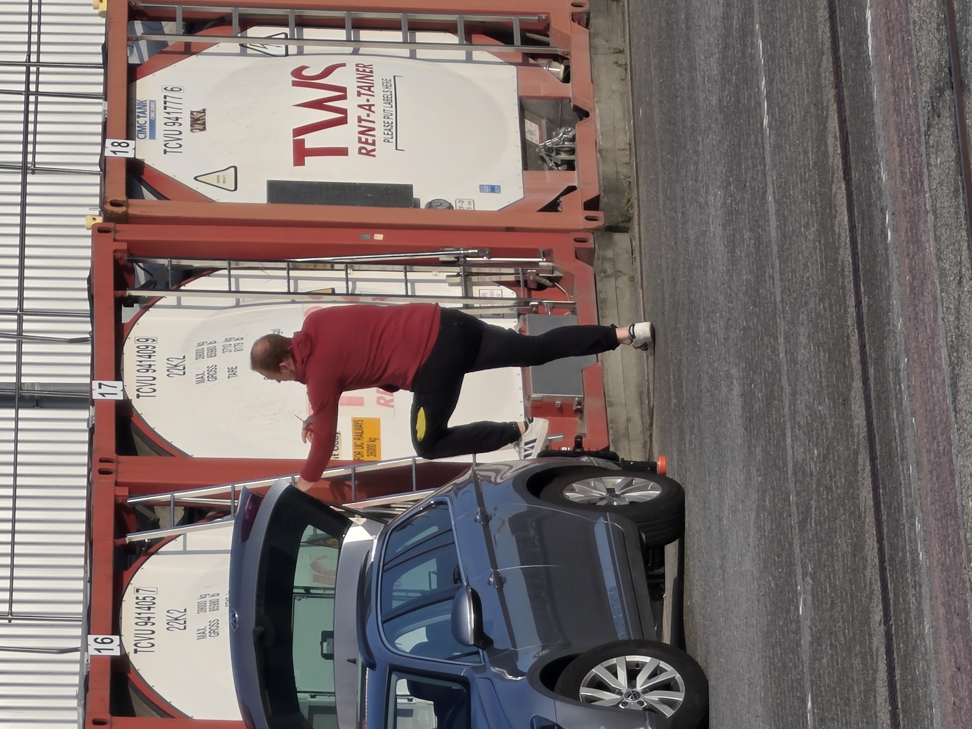 A man in a red hooded sweatshirt and black pants is climbing a ladder attached to a red shipping container. He is facing away from the camera and appears to be reaching for something on the container. A dark gray station wagon is parked next to the container, with its rear hatch open. The scene appears to be an industrial area with multiple shipping containers and a building with a corrugated metal facade in the background. This image was taken in Frankfurt, Germany.