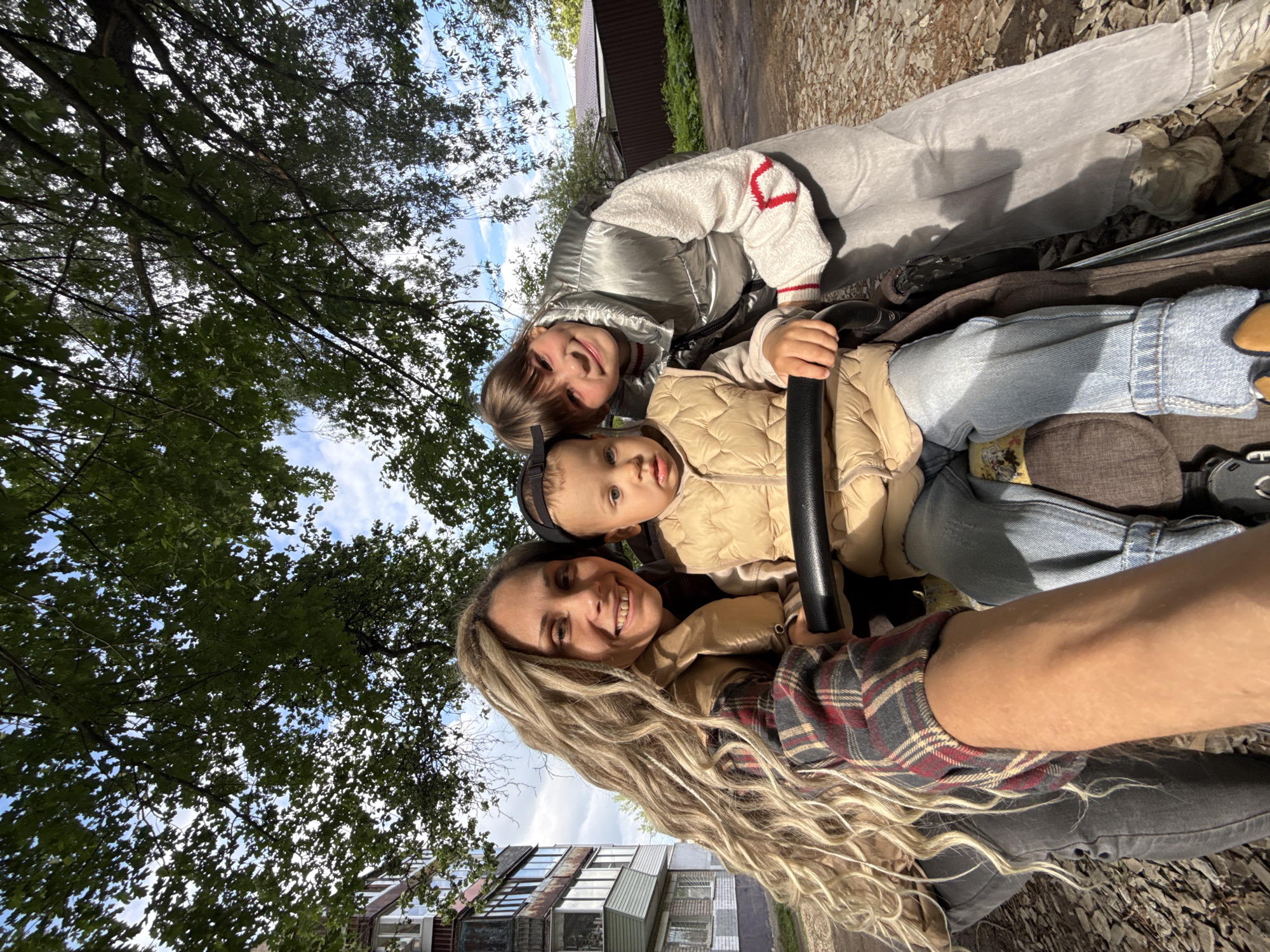 Captured from a low-angle perspective, a selfie shows three individuals in Rusanovo, Russia. A woman with blonde dreadlocks wears a plaid shirt and light-colored jacket. A toddler sits in a stroller, grasping its handle. Behind them is a woman with brown hair wearing a silver jacket with a red accent. The environment includes trees with visible leaves in the background, a building with distinct architecture. The individuals are close together, suggesting a familial relationship.