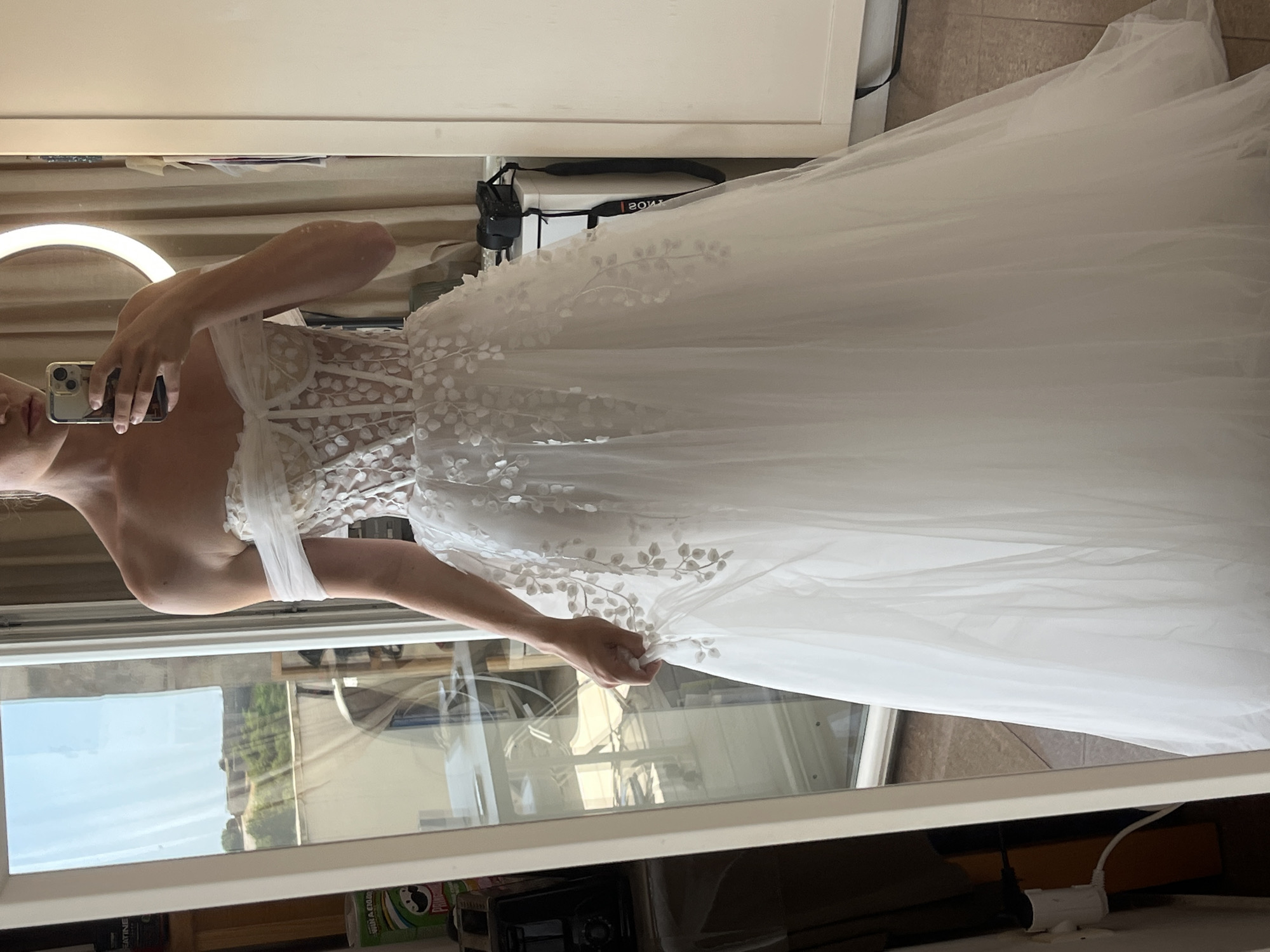 A woman stands in a white, off-the-shoulder wedding dress with a corset bodice and a flowing tulle skirt adorned with floral appliques. She is holding a smartphone to take a mirror selfie. The background includes a large mirror reflecting the room, a doorway leading outside with a view of green hills and a castle, and interior furnishings. The setting appears to be a residential or hotel room in Budva, Montenegro.