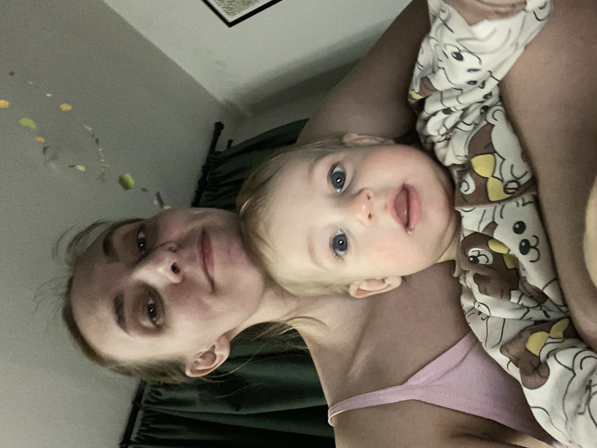 An adult female holds an infant in a close-up, indoor photograph taken in Chaikovsky, Russia. The woman, positioned in the upper half of the frame, has light skin, light brown hair pulled back, and is wearing a light pink spaghetti strap top. She looks directly at the camera with a subtle smile. The infant, held directly in front of the woman, occupies the lower half of the frame. The infant has fair skin, light blonde hair, and wide blue eyes, looking towards the camera with an open mouth. A faint pink flush is visible on the infant's left cheek. The infant wears a long-sleeved garment patterned with cartoon bears. In the background, dark green curtains are visible on the left. The ceiling above is light-colored and features several small, multi-colored decorative pieces. A portion of a dark-framed object is visible against a light wall in the upper right corner.
