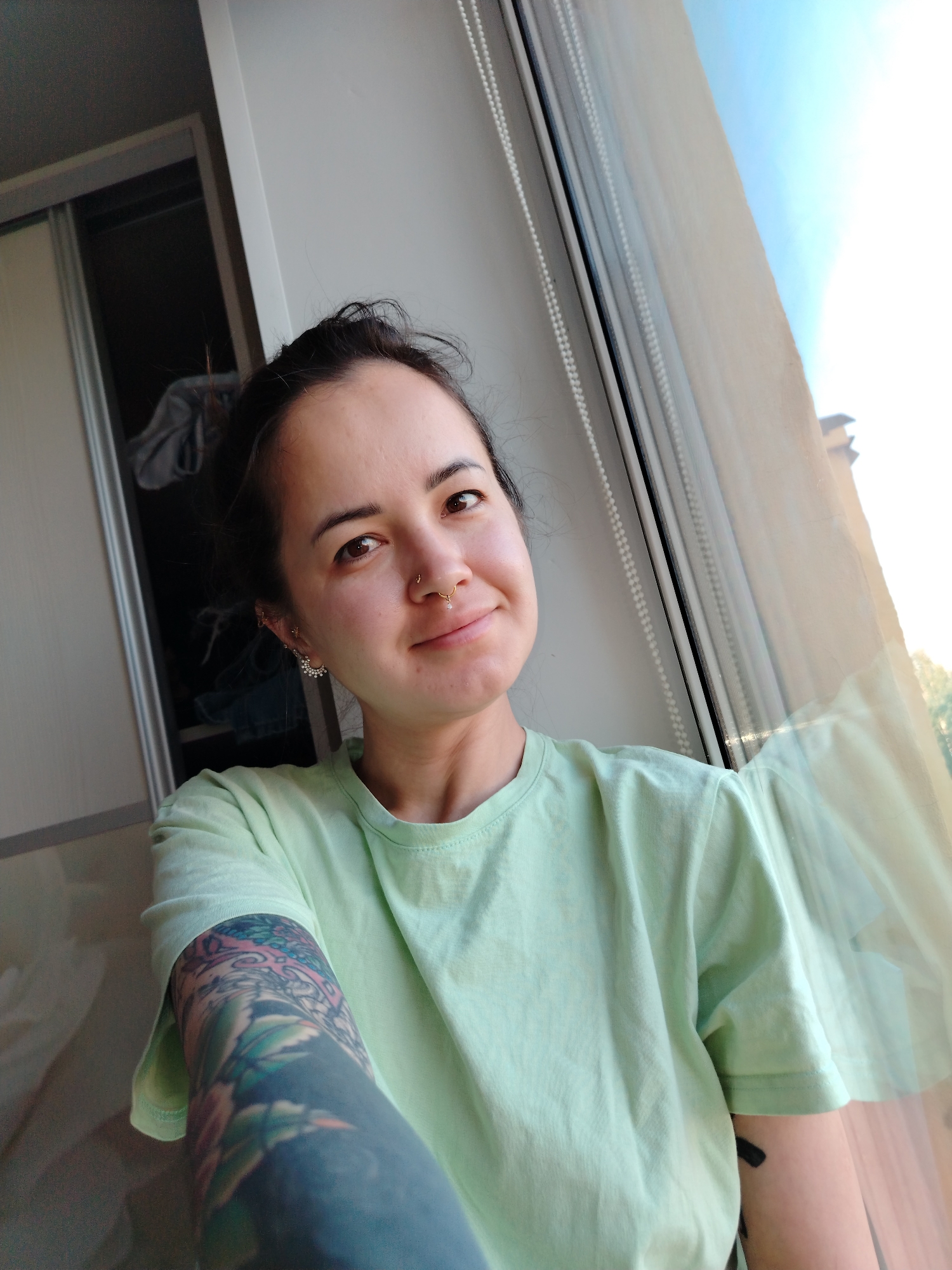 A woman with brown hair pulled back, a nose ring, and multiple earrings is looking at the camera. She is wearing a light green t-shirt and has a full arm tattoo on her left arm. She is positioned near a window with translucent blinds; the blurred cityscape visible outside indicates a residential or urban setting. The background includes a partially open closet revealing clothes inside. The location is Noviy, Russia.