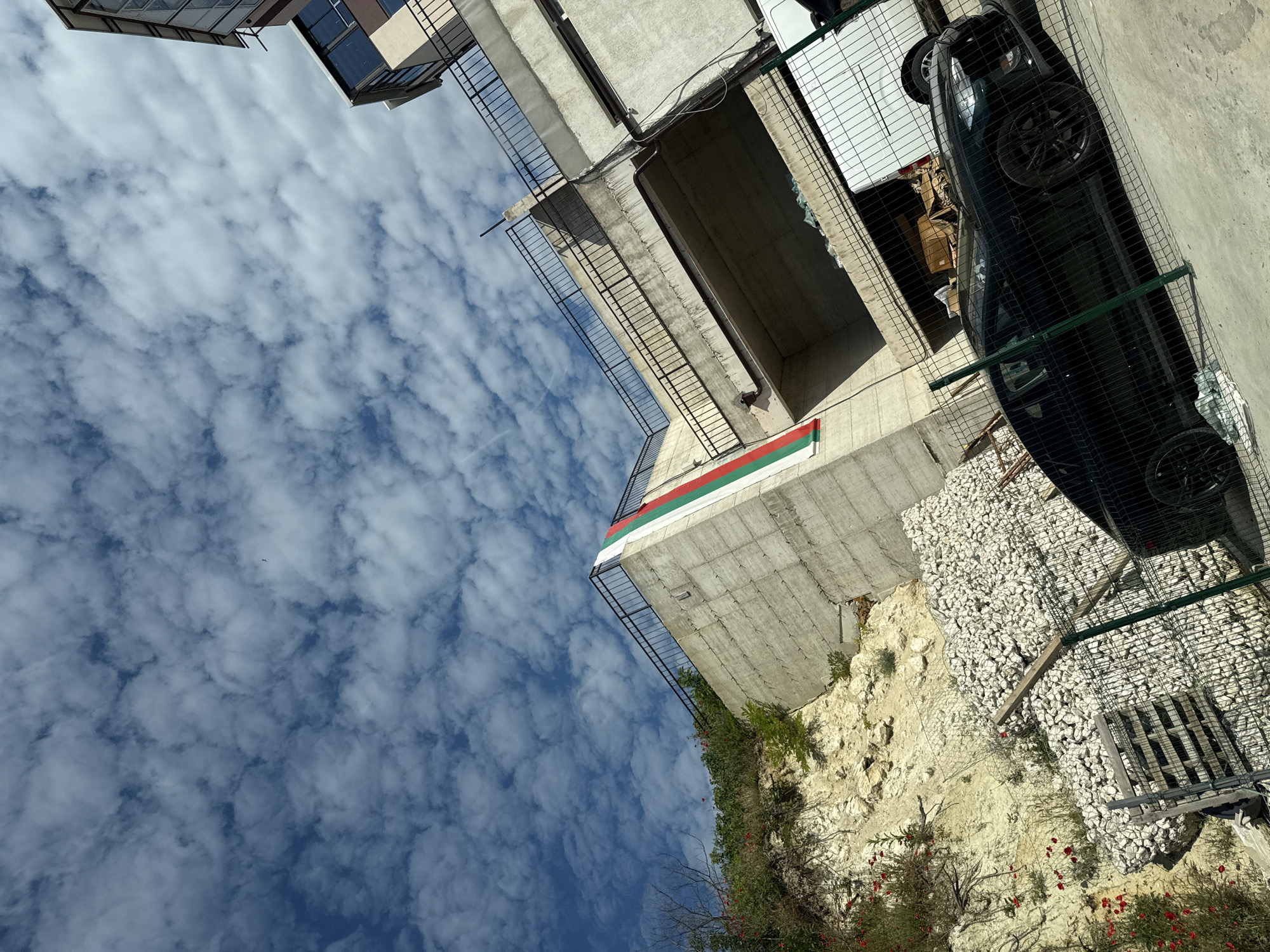The image depicts an outdoor scene with a modern concrete structure, a black sports car, and a white van parked behind a wire fence. A Bulgarian flag is displayed vertically on the side of the concrete structure. The sky is filled with scattered clouds. The foreground shows a section of a wire fence and a black sports car. In the background, the concrete structure rises, with a white van partially visible underneath an elevated section of the structure. The area appears to be an urban environment, possibly a parking lot or an access road. The image is taken from a low angle, looking upwards. There is no visible indication of the specific location within the city of Varna, Bulgaria.