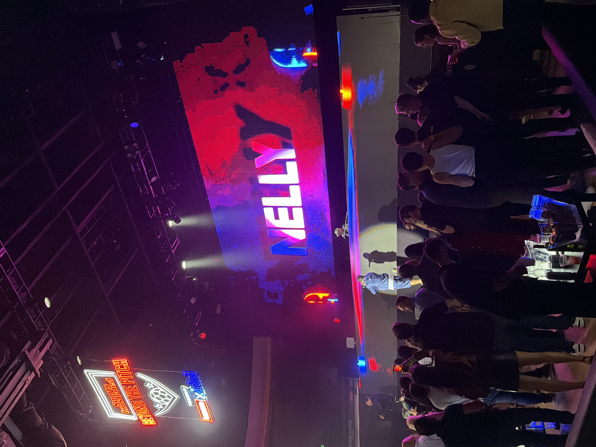 The image depicts an indoor event with a large screen displaying the name "NELLY" in large, colorful letters. Below the screen, a stage or platform is visible with several individuals, including a performer. A crowd of people is gathered in front of the stage, many holding up their phones, suggesting a live performance or concert. Neon signs are visible in the upper left corner, one reading "Sports Illustrated BEYOND THE PITCH." The lighting is dim and colored, with spotlights illuminating the stage. The overall atmosphere appears to be that of a nightclub or concert venue. The location is likely Los Angeles, United States of America, given the context of such events and the presence of known artists like Nelly.