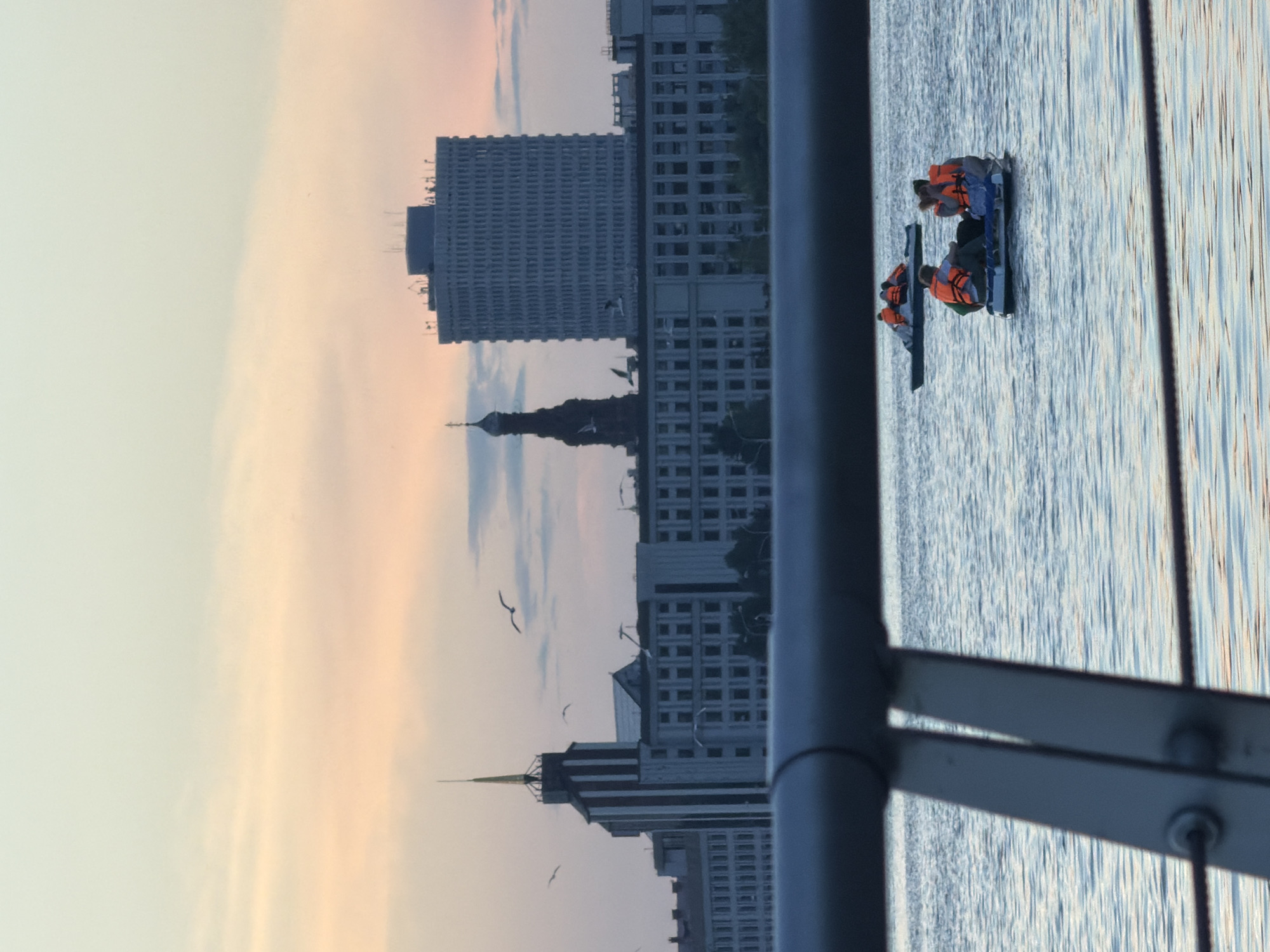 The image depicts a waterscape scene in the city of Kazan, Russia, at either dawn or dusk, characterized by a sky transitioning from light blue-green to soft orange and pink hues. In the immediate foreground, a blurred dark gray metal railing, likely part of a bridge or embankment, extends horizontally across the lower fifth of the frame.
On the body of water, which shows subtle ripples, two small recreational boats are visible. The closer boat, positioned right of center, contains at least four individuals, all wearing bright orange life vests. They appear to be seated and facing each other. A second boat is located further back and to the left, also carrying multiple individuals in orange life vests.
The background reveals the Kazan city skyline. To the left, a prominent building with a tall, slender, dark spire and vertical lighter stripes is visible, identifiable as a significant structure in Kazan, possibly related to its educational institutions (e.g., Kazan State Technical University). To the right of this, a distinctive dark, spired structure, possibly a historic tower or a religious building (like the Söyembikä Tower or a church bell tower), stands against the sky. Further to the right, a large, rectangular, multi-story building with numerous small, uniform windows dominates the mid-right background, resembling a typical administrative or office block. Various lower-rise buildings complete the urban backdrop. Several seagulls are observed flying against the sky, particularly above the buildings.