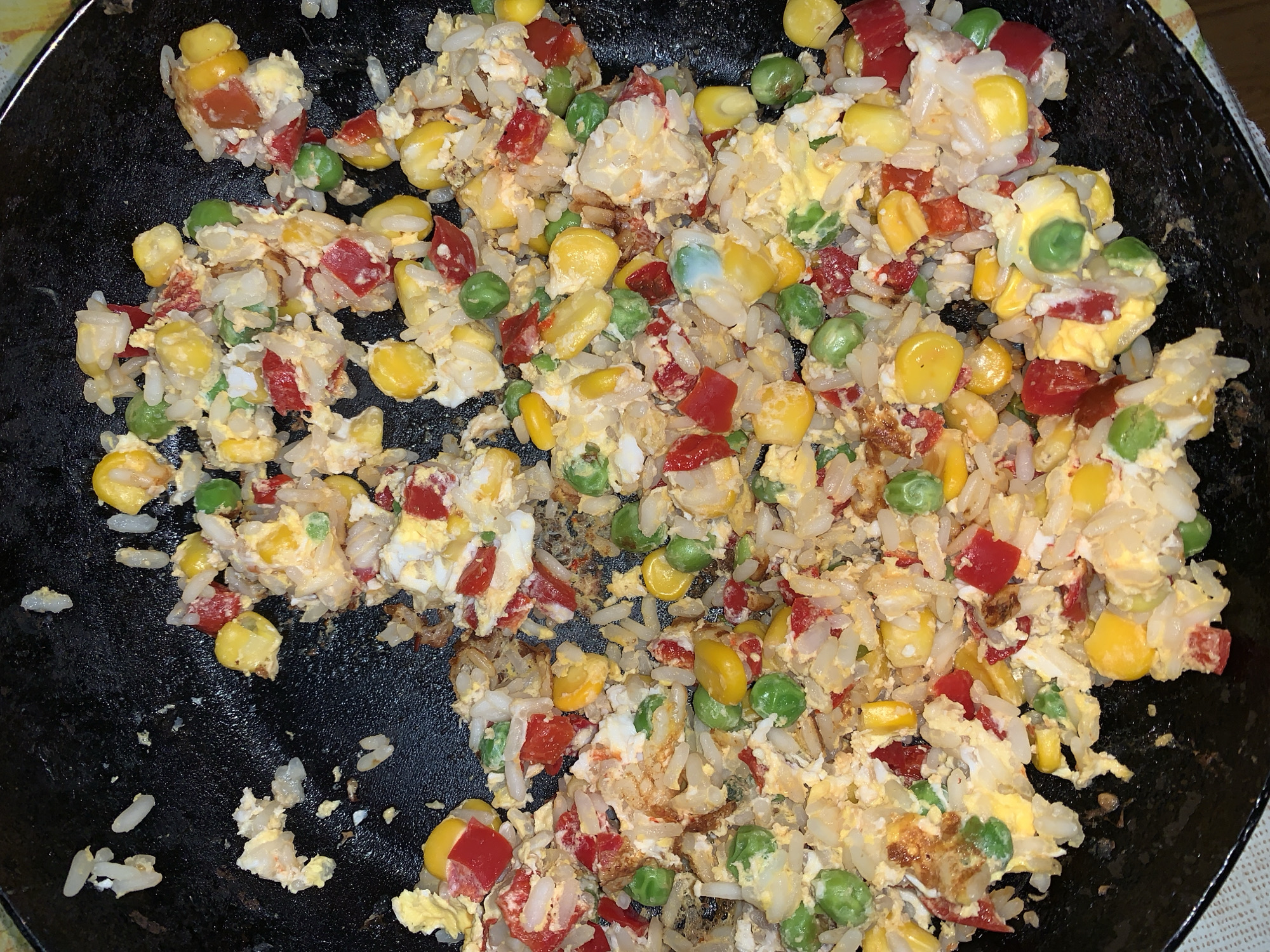 The image shows a close-up of fried rice in a black frying pan. The ingredients include white rice, scrambled eggs, corn, peas, and diced red bell peppers. The dish appears to be freshly cooked and is still in the pan.