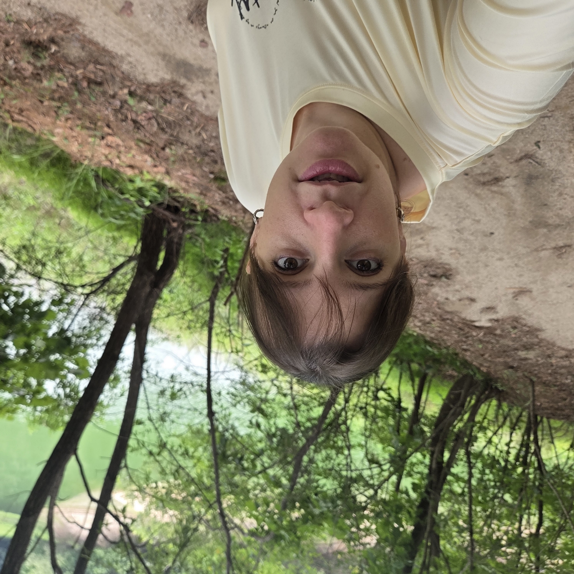 In Gapcheon-myeon, South Korea, a woman takes a selfie on a dirt path in a wooded area. She is wearing a pale yellow t-shirt and hoop earrings. The background features lush green trees, and a body of green water is visible through the foliage. The lighting suggests an overcast or shaded day.