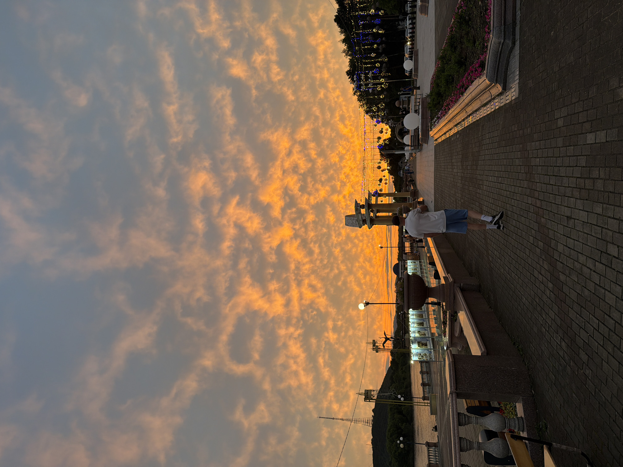 The image depicts a sunset over the Amur River embankment in Birobidzhan, Russia. A man wearing a white t-shirt and blue shorts is walking away from the camera on a brick-paved walkway. The sky is filled with dramatic orange and yellow clouds. Along the walkway, there are stone balustrades, lampposts, flowerbeds, and decorative spherical lights strung between trees. In the background, a radio tower is visible on a hill, and the river stretches out to the horizon. The overall scene is a picturesque urban landscape bathed in the warm glow of the setting sun.