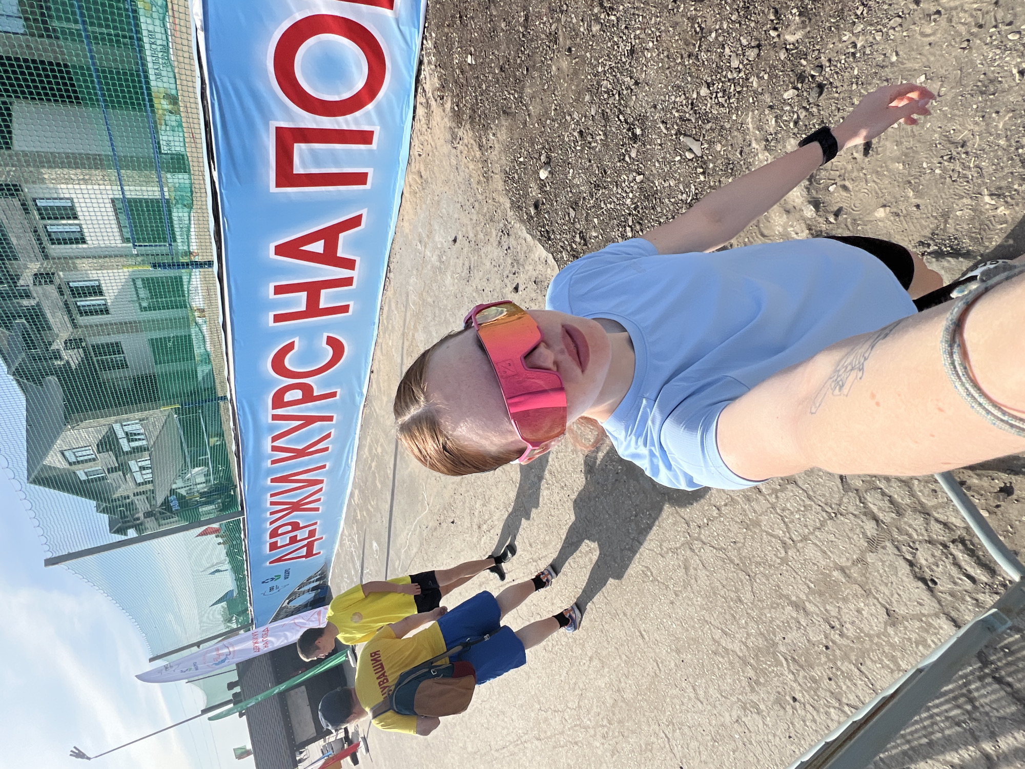 A young woman takes a selfie at what appears to be an outdoor sports event, possibly a race, in Khvalynsk, Russia. She is wearing a light blue t-shirt and pink sunglasses. In the background, two men in yellow t-shirts and blue shorts are standing nearby. A blue banner with red Cyrillic text "ДЕРЖИ КУРС НА ПО..." is visible. A mesh fence and a building can be seen in the background, suggesting a sports facility. The ground is a mix of concrete and gravel.