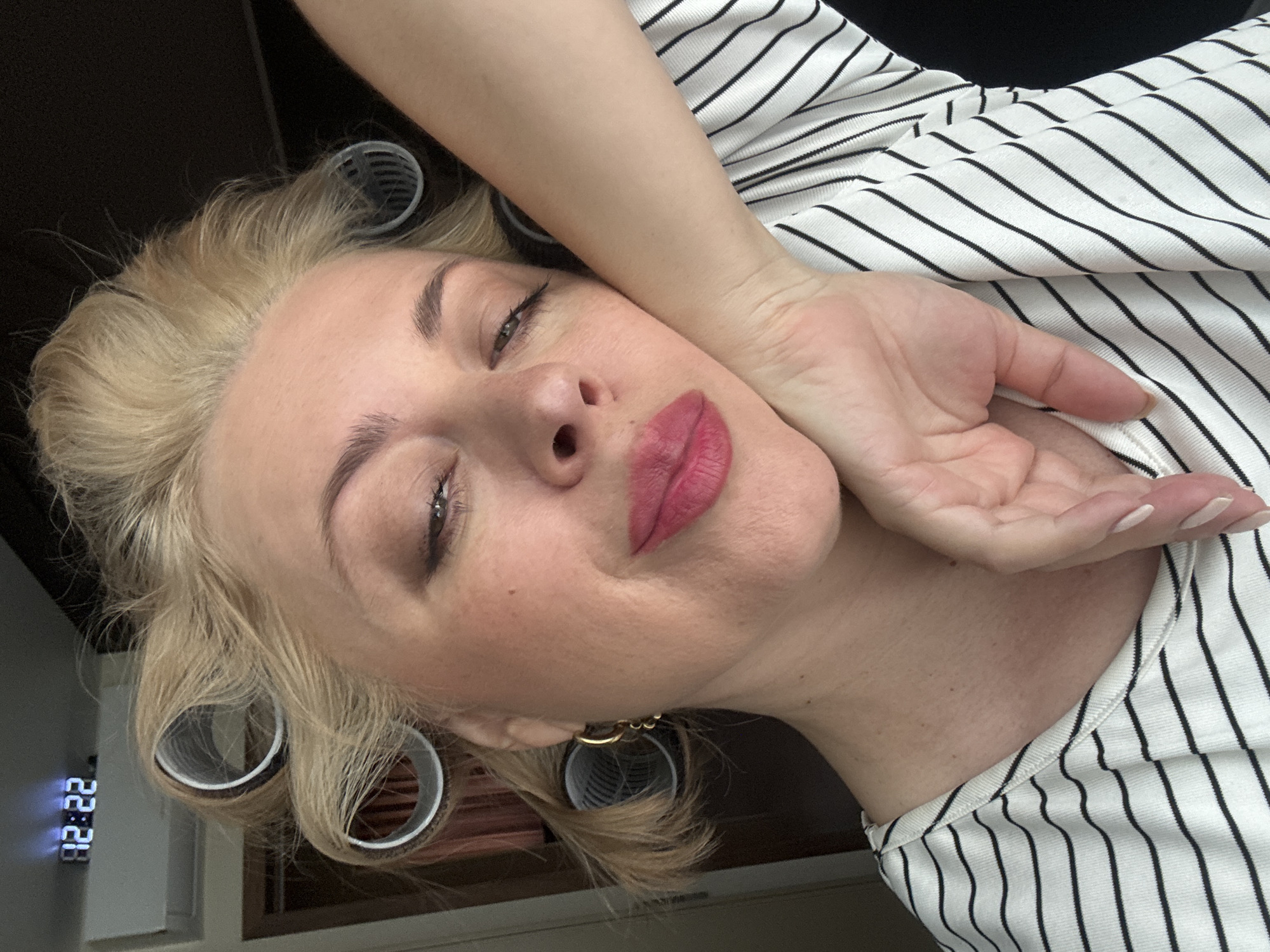 A woman with blonde hair styled in rollers poses for a selfie. She is wearing a black and white striped shirt and has bright pink lipstick on. Her left hand is resting on her cheek. In the background, a digital clock displays "12:22". The scene appears to be indoors, possibly in a bedroom, as suggested by the presence of hair rollers and the clock. The overall impression is a casual, at-home moment. There is no indication of the city of Sochi, Russia.