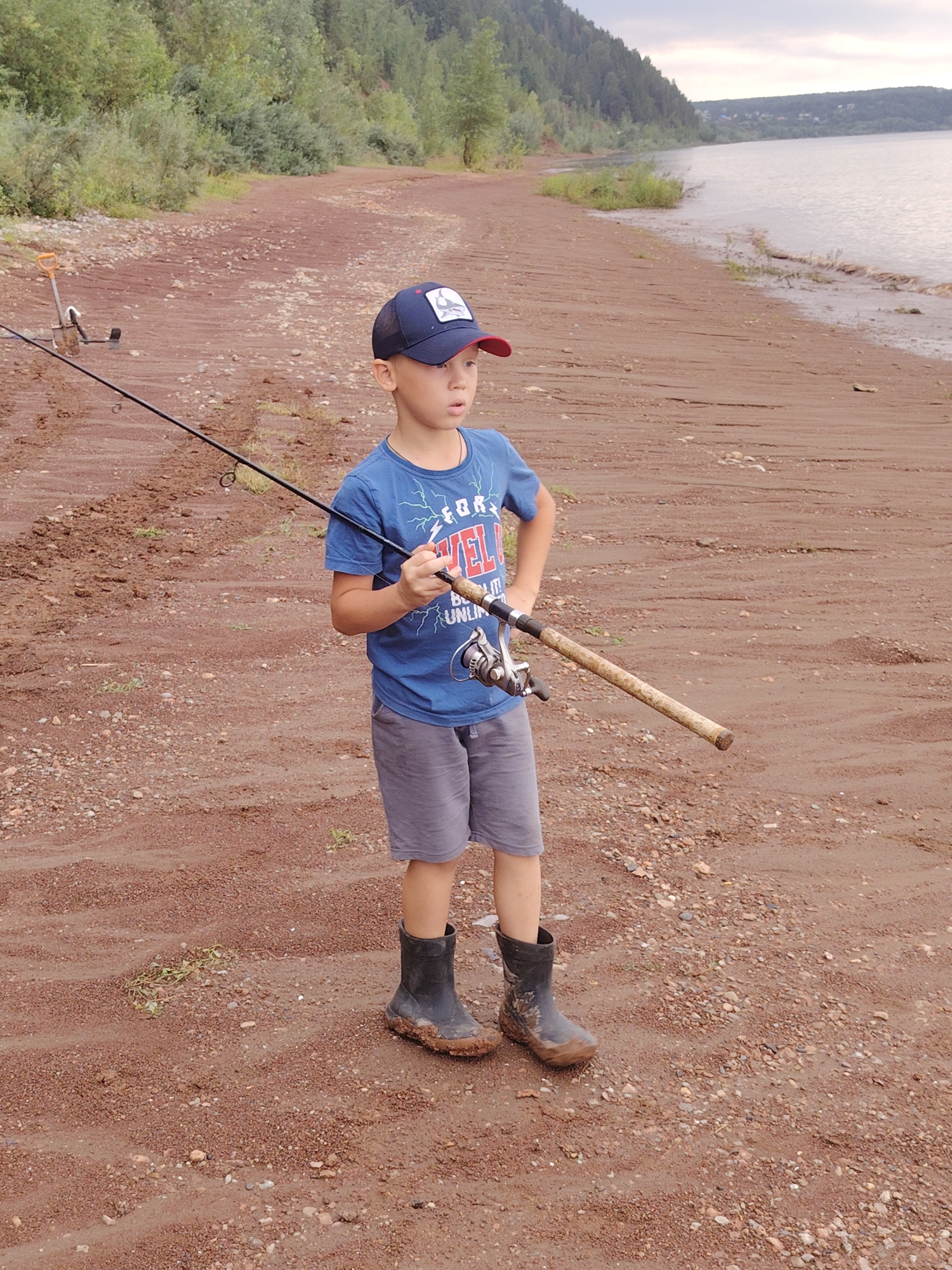 A young boy wearing a blue t-shirt, grey shorts, a navy blue baseball cap with a graphic, and black rubber boots stands on a reddish-brown sandy beach holding a fishing rod. The fishing rod is a dark color with a silver reel and a light brown wooden handle. The boy is looking to his right, with his mouth slightly open. Behind him, the beach stretches towards a body of water, likely a river, which is calm and reflects the overcast sky. The shoreline is lined with green trees and a forested hill. In the foreground, the sandy ground is textured with small pebbles and some scattered dry grass. A fishing rod holder is visible in the background to the left of the boy. The overall environment suggests a rural or natural setting, possibly on the outskirts of a city.