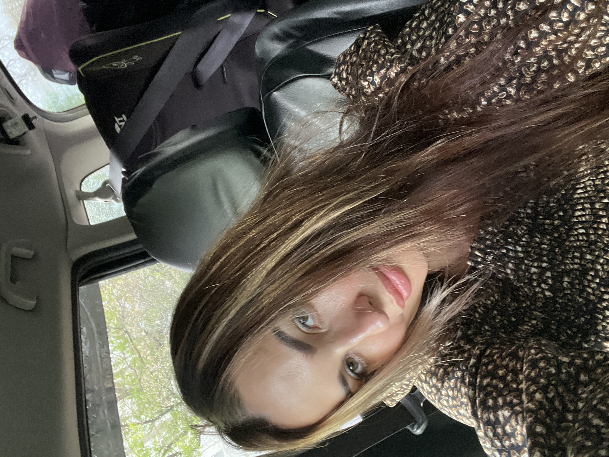 A woman is taking a selfie in a car. She is in the passenger seat, with a child seat visible behind her. The woman has long, wavy, brown hair with blonde highlights. She is wearing a leopard print jacket. The interior of the car is dark, with the outside visible through the windows. It appears to be raining outside. The location is not specified.
