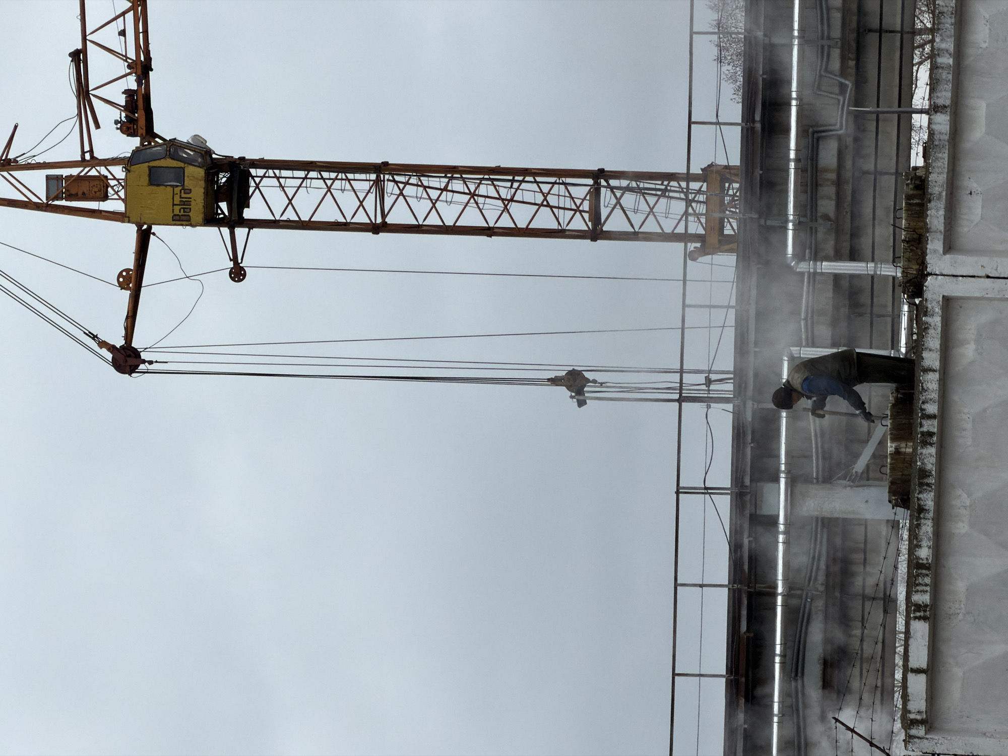 The photograph depicts a city scene with a focus on industrial elements. In the upper left quadrant, a tall, weathered, orange-yellow crane dominates the view against a dull, overcast sky. The crane is a lattice structure, and its control cabin bears the inscription "Bakira." Running horizontally across the frame in the middle-right portion of the picture, a dilapidated, concrete structure is visible, including a network of pipes. A worker, dressed in muted colors and wearing a beanie, can be seen leaning over the edge of the structure, appearing to perform some sort of maintenance. The scene is further complicated by the presence of mist or steam emanating from the vicinity of the pipes, obscuring the view somewhat. The presence of a barbed wire fence at the base of the structure adds to the industrial feel of the image. Based on the metadata and tags, the city is Chaikovsky, Russia.