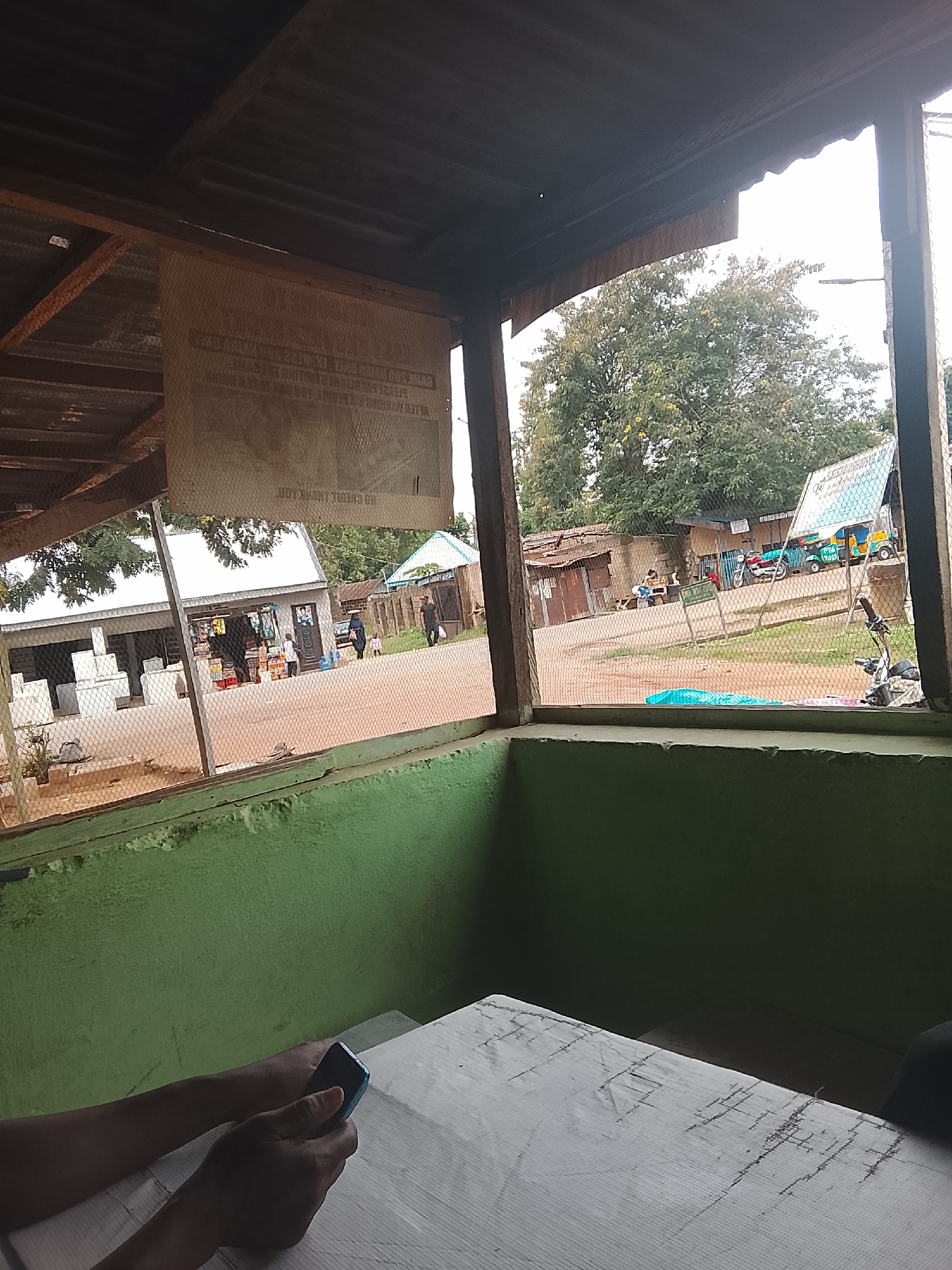The image depicts an outdoor scene, viewed from under a rustic wooden shelter with a corrugated metal roof. In the foreground, a person's hands are visible holding a dark blue mobile phone, resting on a white, textured table. The table appears to have some marks or damage.
Through the opening of the shelter, a street scene unfolds. Several buildings, including what appears to be a small shop with goods displayed outside, are visible. People are milling around the area, some seemingly interacting at the shop, while others walk by. A few vehicles are also present, including a motorcycle and a bright yellow and blue auto-rickshaw. A white, fenced enclosure with what look like concrete structures is also part of the scene.
In the background, lush green trees dominate the landscape, suggesting a somewhat rural or suburban setting. The sky is overcast, hinting at a cloudy or possibly humid day. The overall impression is of a casual, everyday scene in a community. A large, light-colored sign with text, some of which appears to be in a foreign language and is partially obscured by a mesh fence, hangs from the shelter. The text on the sign is not clearly legible. The atmosphere appears calm and ordinary.