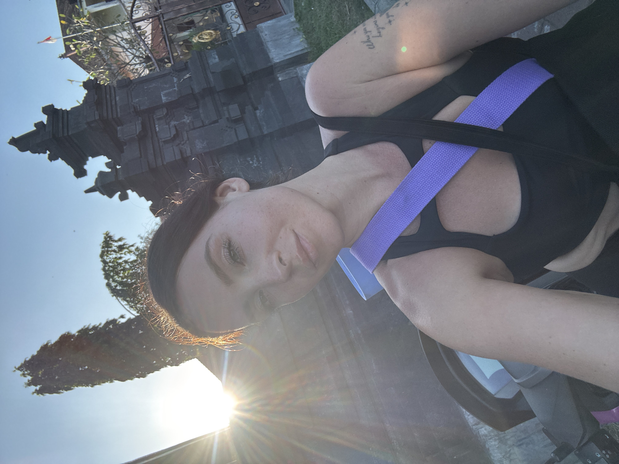 A medium shot captures a woman from the shoulders up in a selfie taken outdoors in North Kuta, Indonesia. The woman is in focus, with the sun setting behind her, creating a lens flare that extends across the left side of the image. She has dark hair pulled back and is wearing a black athletic top with a purple strap across her chest. A tattoo is visible on her right arm. In the background, a dark, ornate stone structure resembling a temple gate stands to the right, with a red and white flag and a building behind it. A tall, slender tree is visible on the left, silhouetted against the bright sky. The lighting suggests it is late afternoon or early evening.