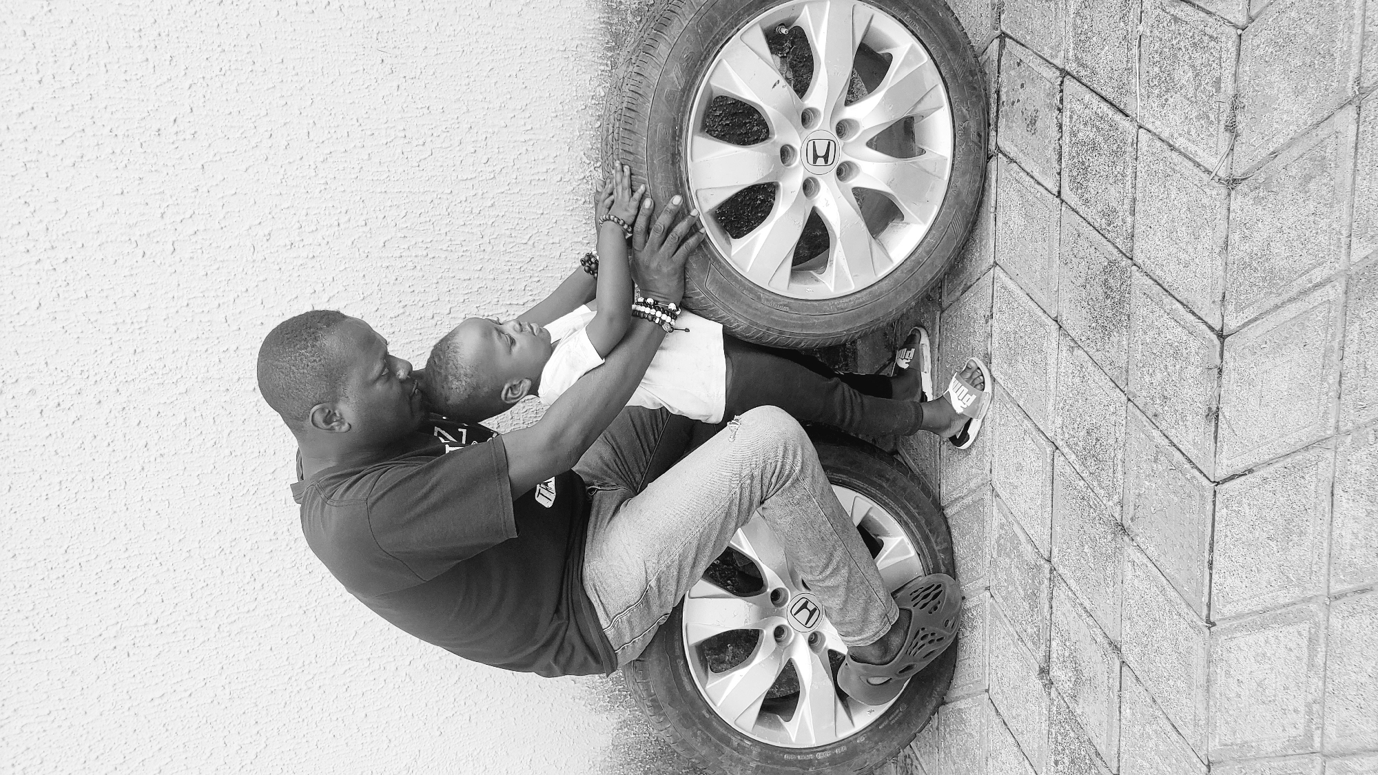 This black and white photograph captures a tender moment between an adult and a child in Narayi, Nigeria. The image is oriented with the subjects lying horizontally across the frame, against a textured white wall and patterned concrete ground.
**Subjects:** The primary subjects are an adult male and a young child. The adult, wearing a dark t-shirt, light jeans, and dark slip-on shoes, is seated on a car tire. He is holding the child, who is dressed in a white t-shirt, dark pants, and white sandals with "PUMA" text visible. Both individuals wear beaded bracelets on their wrists.
**Activity or event taking place:** The adult is embracing the child, leaning in to kiss or nuzzle the child's head/face. The child is lying back against another car tire, with arms extended upwards, resting on it. The scene portrays a loving and affectionate interaction.
**Notable details:** Two car tires, each featuring a prominent Honda logo on its rim, are central objects, providing a backdrop and seating for the subjects. The setting appears to be an outdoor or semi-outdoor space, characterized by the rough-textured white wall and the geometric pattern of the concrete pavement. The lighting suggests daylight, creating soft shadows. The overall emotion conveyed is one of warmth, parental love, and comfort.