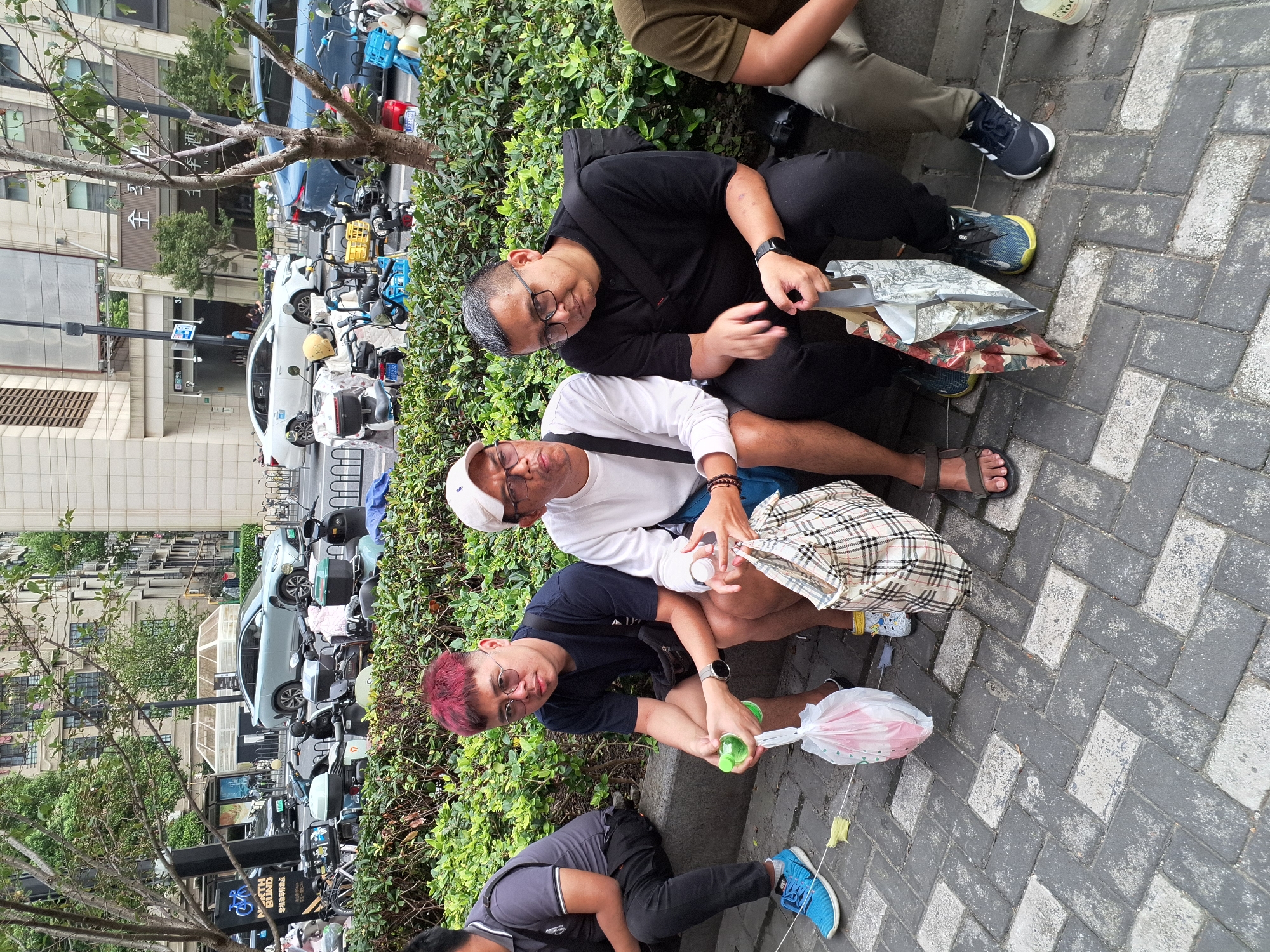 Three men are sitting on a raised concrete curb in front of a dense green hedge. The man on the left wears a dark grey t-shirt and black pants, with bright blue sneakers. He holds a green water bottle and a white plastic bag. The middle man has pink-dyed hair, wears a dark blue t-shirt and black shorts, and also has bright blue sneakers. He is holding a white plastic bag. The man on the right wears a black t-shirt and black pants, with black and blue sneakers. He has a black bag slung across his body and is holding a patterned reusable shopping bag. Behind the men, there is a street with several parked electric scooters and cars. In the background, tall buildings are visible. The scene appears to be in an urban environment, likely Hongkou District, China, given the presence of the parked scooters and the architectural style of the buildings. The pavement is made of interlocking grey and white rectangular stones.