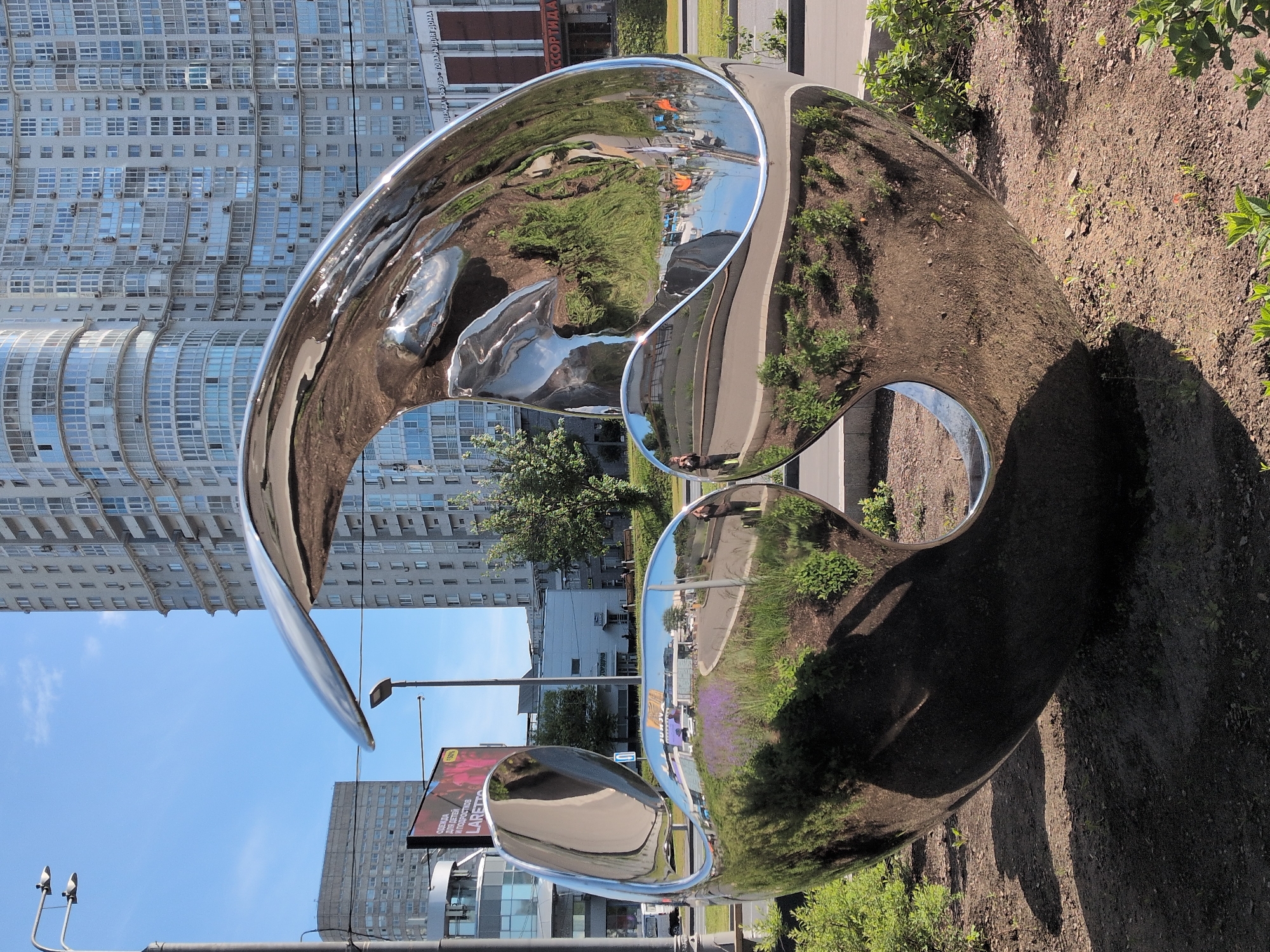 A large, polished metal sculpture is positioned on a strip of land between a sidewalk and a street, likely in Moscow, Russia. The sculpture reflects the surrounding urban environment, including buildings, trees, and people. The sculpture's shape consists of curves and openings, creating a distorted reflection of the area. The ground surrounding the sculpture is covered with soil and vegetation. In the background, a large building with numerous windows is visible, suggesting a densely populated urban setting. A street with vehicles and streetlights further indicates the city's character.