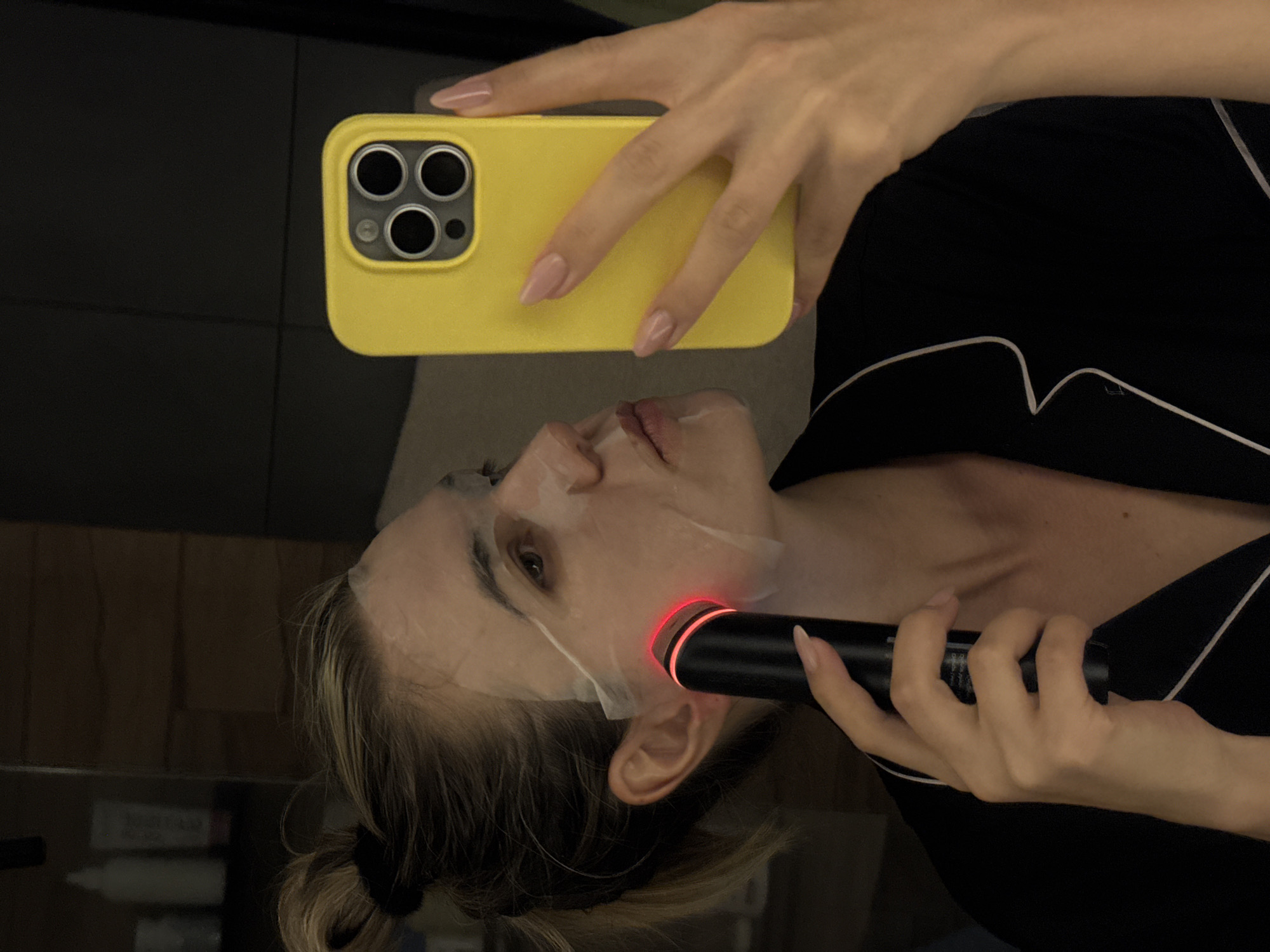 A woman with blonde hair pulled back in a low ponytail is holding a black cylindrical device with a red light against the right side of her face. She has a white sheet mask on her face. In her left hand, she holds a yellow iPhone with three camera lenses, taking a selfie. She is wearing a black top with white trim. The background is dark with reflective surfaces.