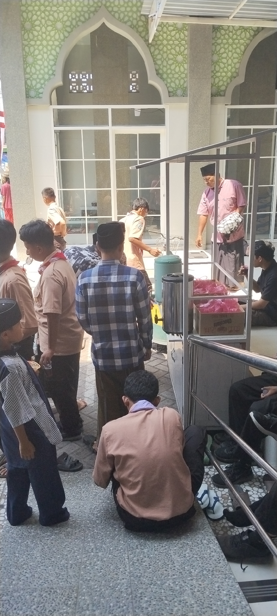 In Jombang, Indonesia, a group of people, predominantly young males in uniforms resembling scouts or similar organizations, are gathered outside a building with Islamic architectural elements, possibly a mosque or a related religious institution. The building features arched windows with decorative green geometric patterns.
Several individuals are dressed in light brown uniforms with red scarves or neckerchiefs. Some are standing, while others are seated on the ground. One person in the foreground, seen from behind, wears a blue and white checkered long-sleeved shirt and brown pants, with a light purple scarf around his neck. Another young person, also seen from behind and to the left, wears a dark blue and white striped tunic over dark pants.
In the background, an adult male wearing a pink shirt and a peci (a traditional cap) is interacting with a young person in uniform. They are standing near a makeshift food or drink stall, which has a metal frame and shelves displaying pink plastic cups and a box labeled "Chocolate." There are also large green and silver containers, possibly for water or other beverages, near the stall.
The ground is paved with textured grey tiles. Sunlight is casting shadows, indicating an outdoor or semi-outdoor setting. The overall atmosphere appears to be one of community gathering or an event.