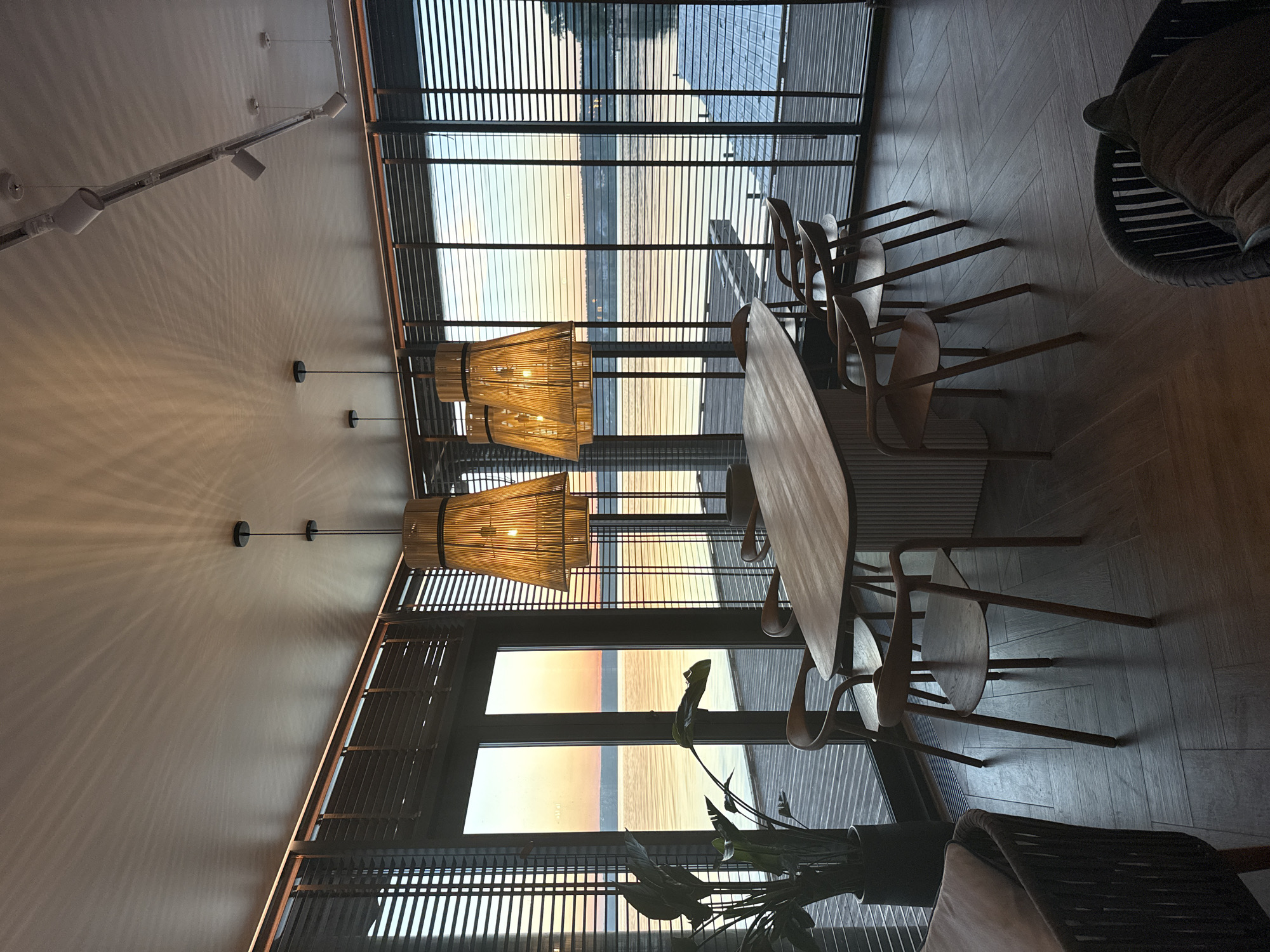An interior photograph taken in Votkinsk, Russia, depicts a contemporary dining or lounge area with a expansive view of a large body of water during twilight hours, likely sunrise or sunset. The ceiling, painted a light, uniform color, is equipped with several lighting elements: three prominent, bell-shaped pendant lamps featuring woven, possibly rattan, shades are suspended over a rectangular wooden dining table. These lamps emit a warm glow, casting distinct, elongated, wavy shadow patterns across the ceiling surface. A recessed linear light strip is installed along the top edge of the window framing, and a track lighting system with three dark, cylindrical spotlights is mounted on the upper right portion of the ceiling.
The exterior wall of the room consists of large, dark-framed, floor-to-ceiling windows, each fitted with dark horizontal slatted blinds that are partially open. Through these windows, a tranquil body of water, consistent with the Votkinsk Reservoir, is visible, reflecting the pastel hues of the sky: a gradient of light blue, soft orange, yellow, and pink near the horizon. A wooden deck or pier is present immediately outside the windows, extending over the water. A distant shoreline or landmass is faintly discernible across the reservoir.
The interior furnishings include a long, light-toned wooden dining table with rounded ends and a vertically fluted or ribbed base. Positioned around the table are at least six minimalist wooden chairs, characterized by curved backrests and seats. In the lower left quadrant of the image, a portion of a dark, woven armchair containing a light-colored cushion is visible. Similarly, in the lower right, another dark, woven armchair with a dark cushion is partially present. A tall, dark-potted plant with large, glossy green leaves stands next to the leftmost window. The floor is composed of dark-colored tiles arranged in a herringbone pattern. No individuals are present in the scene.