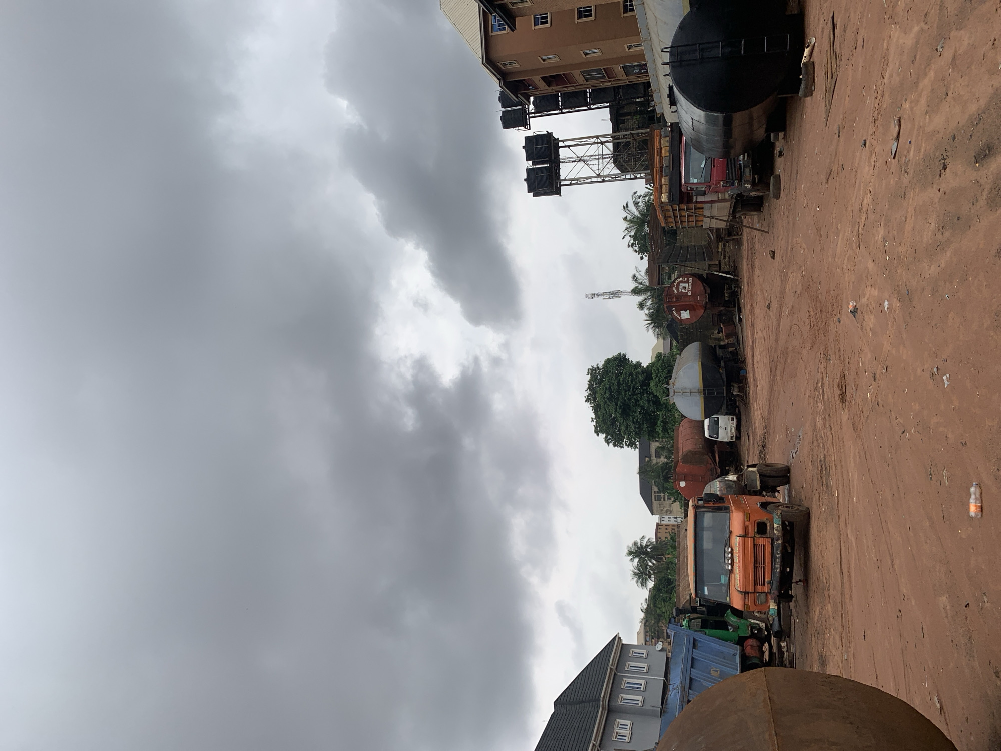 The image captures a dusty, outdoor scene in the city of Nkpor, Nigeria, likely a truck depot or repair yard. The ground is a reddish-brown dirt, with scattered debris including plastic bottles and discarded items. In the foreground, a large, curved metal object, possibly a storage tank, takes up the left side of the frame. Behind it, various trucks and tankers are parked.
Dominating the center is an orange tanker truck with a front-mounted engine. To its left is a blue dump truck. Further back, several other tanker trucks are visible, some red, some black, and at least one with a white cab. One red tanker has "HIGHLY INFLAMMABLE NO SMOKING" written on it. In the background, to the right, a multi-story residential building with a light brown facade and multiple windows stands. To its left, a metal tower supports several large black water tanks. Behind the buildings and trucks, lush green trees and palm trees suggest a tropical climate. The sky above is overcast with heavy, gray clouds, indicating a potentially rainy day. There are no people or animals clearly visible in the image.