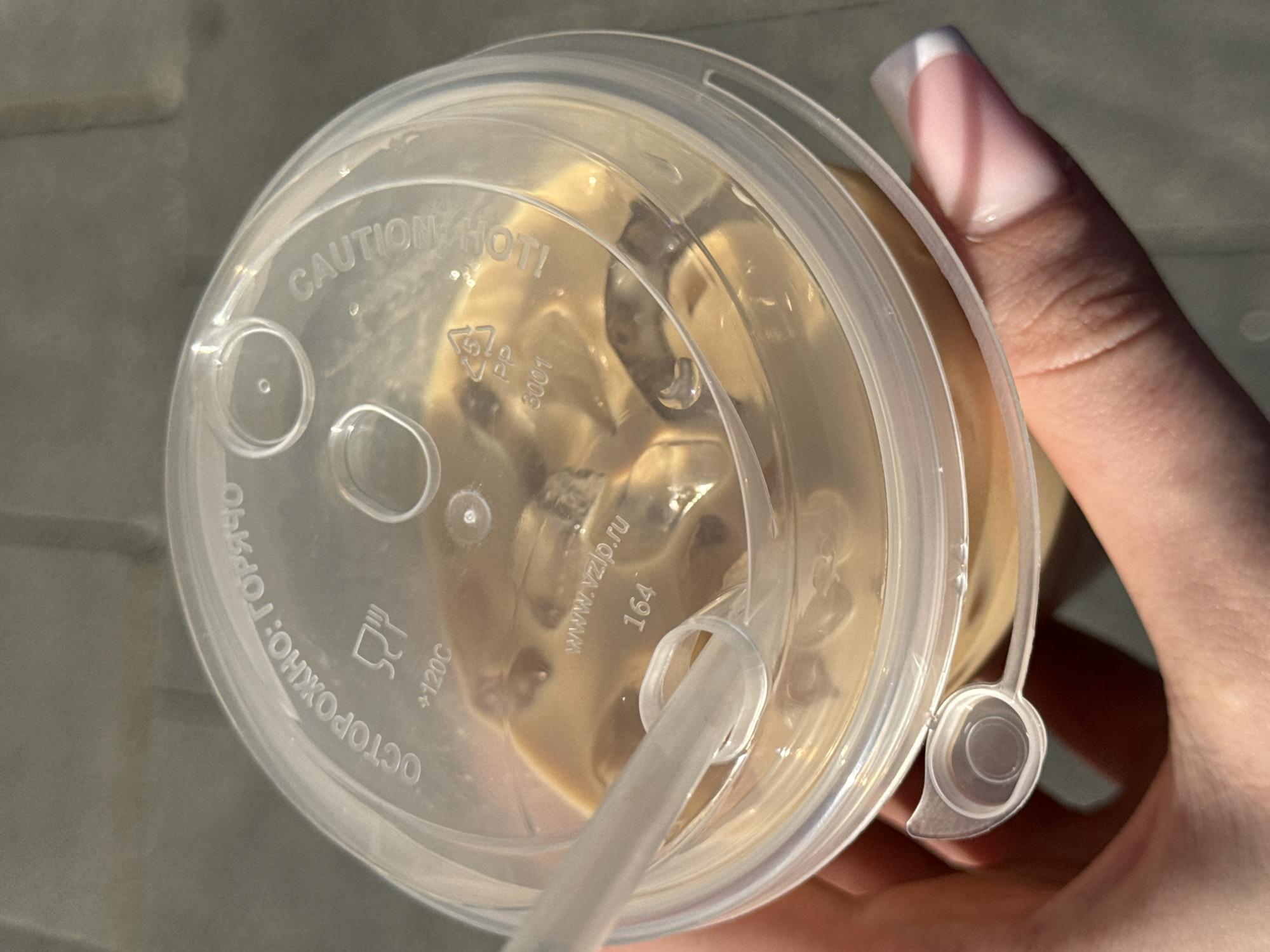 A close-up, top-down shot shows a clear plastic cup filled with a light brown, iced beverage, held by a person's hand. The cup is topped with a clear plastic lid that has embossed text. The text in Cyrillic on the lid reads "ОСТОРОЖНО: ГОРЯЧО" which translates to "CAUTION: HOT." Below this, there is a recycling symbol for "PP" (polypropylene) and the number "3001". Further to the right, the word "CAUTION" is visible, also embossed on the lid. The beverage appears to be coffee with milk or a similar drink, with ice cubes visible within. A white plastic straw is inserted into the lid. The person's hand, which is visible at the bottom and left of the frame, has clean, manicured fingernails with a French manicure. The background appears to be a gray, paved surface, possibly an outdoor area in a city. The lighting suggests it is daytime, with sunlight casting some shadows. The image does not contain any recognizable landmarks or specific details that would indicate it is located in Moscow, Russia.