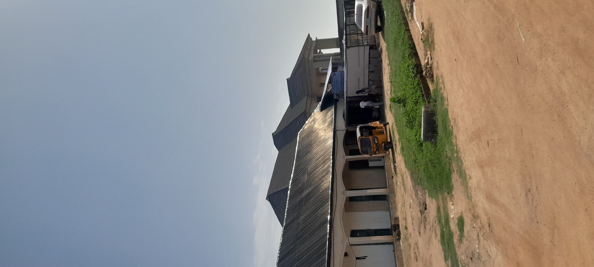 A wide shot shows a building with a corrugated metal roof extending from the left to the center, with another building with a darker tiled roof behind it, and a portion of a larger structure with columns in the background. A yellow auto-rickshaw is parked in front of the corrugated-roof building, next to a white car. Two people, one wearing a white shirt, stand near the auto-rickshaw. The ground is unpaved, with patches of dry grass and some scattered debris. The sky is clear and light blue. The scene appears to be located in an outdoor area, possibly a commercial or residential space, in Jalingo, Nigeria.