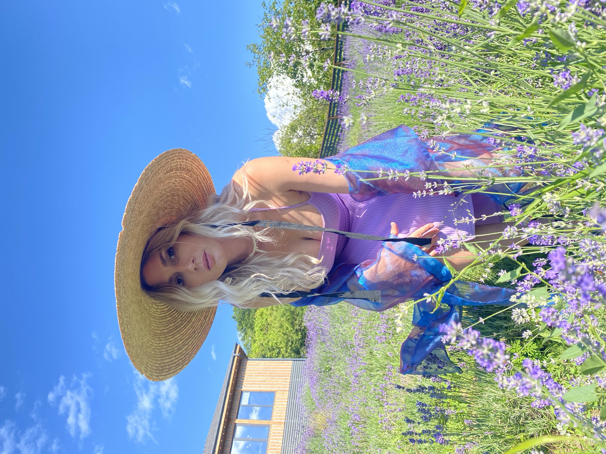 A woman with blonde hair wearing a large straw hat, a purple dress, and a translucent blue and pink overcoat kneels in a field of lavender. She is looking at the camera. To her left, a modern wooden building with large windows is visible. The sky is blue with scattered clouds. The scene appears to be a lavender farm in Verkhneye Uchdere, Russia.