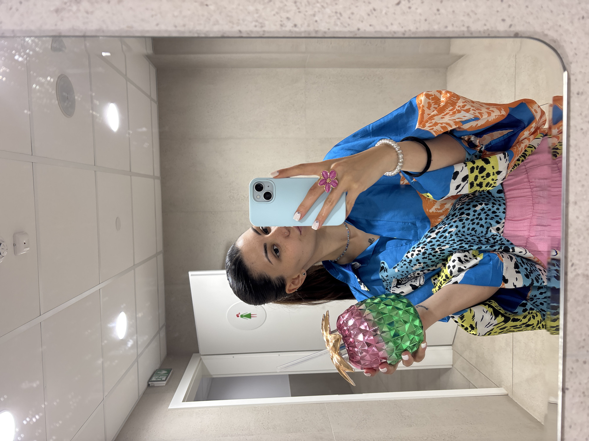 A woman is taking a selfie in a restroom in Sofia, Bulgaria. She is holding a cell phone in her left hand and a pineapple-shaped object with a gold top in her right hand. The restroom has white walls, white doors, and a grey floor. The woman is wearing a blue shirt with an animal print and a pink skirt. She is wearing a bracelet, a ring, and a necklace. The restroom contains a door with a green sign depicting a woman wearing a dress.