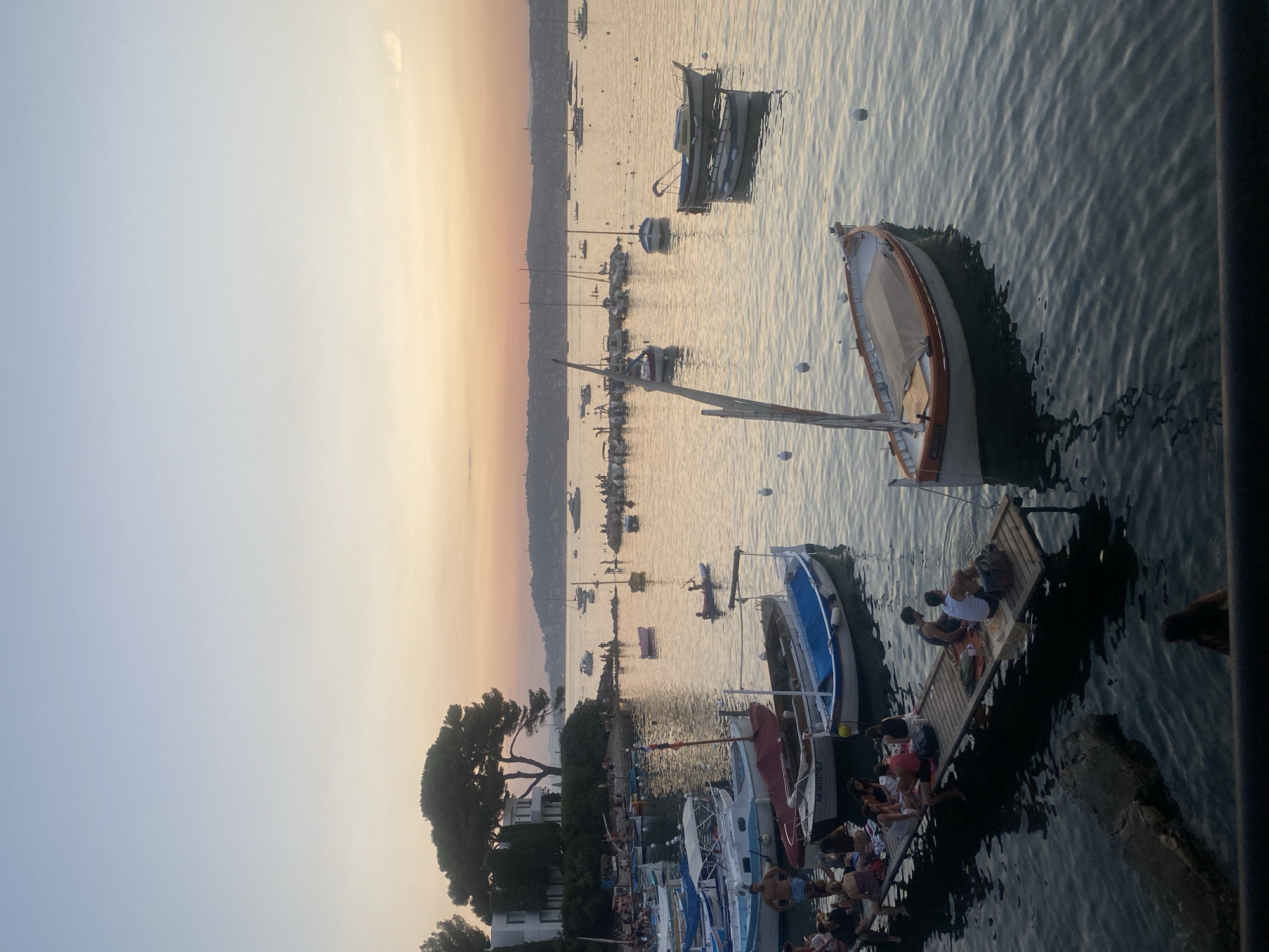 The image depicts a harbor scene in Antibes, France, at dusk. Numerous boats, varying in size and style, are moored in the calm water. Some are small, weathered fishing boats, while others appear to be yachts. Several people are visible on a wooden pier in the foreground, some seated, others standing, and a few are in the water. In the background, a tree-lined coastline with buildings suggests a developed area. The sky exhibits a soft gradient of blue and orange, indicative of sunset. The overall atmosphere is serene and leisurely.